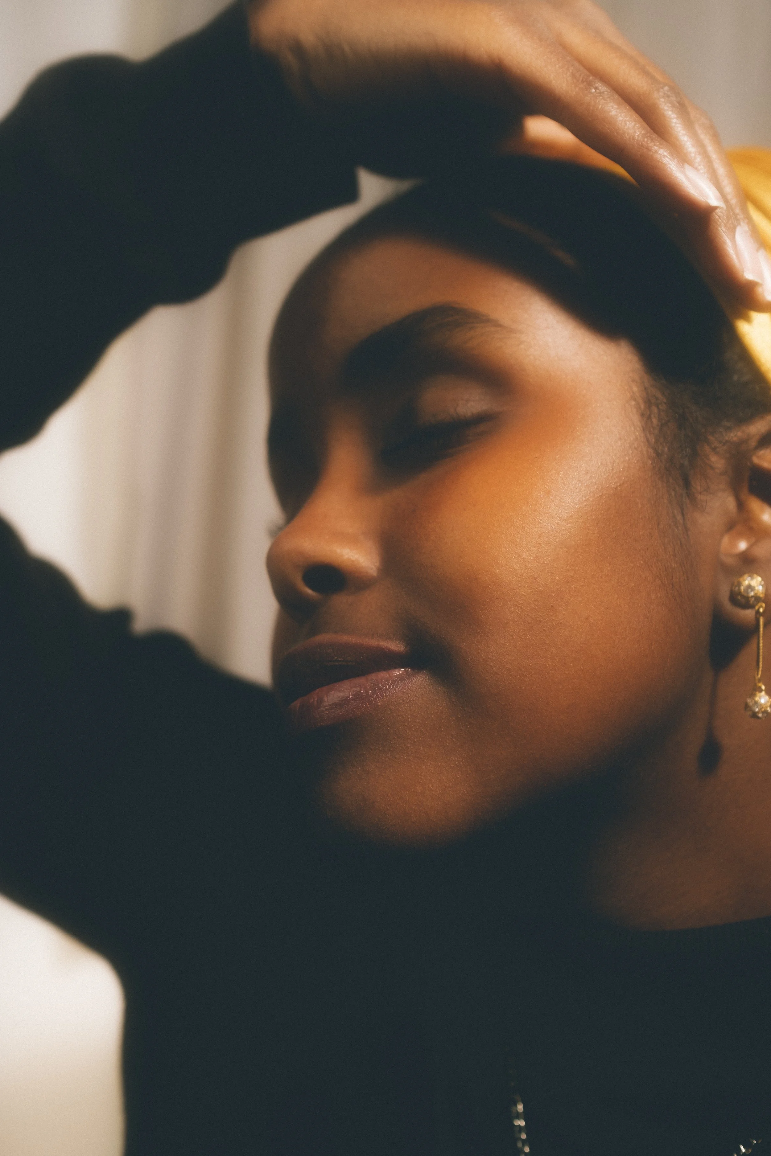 A close-up portrait of a woman with her eyes closed, wearing gold earrings and a black long-sleeve top, touching her forehead with one hand, and smiling softly.