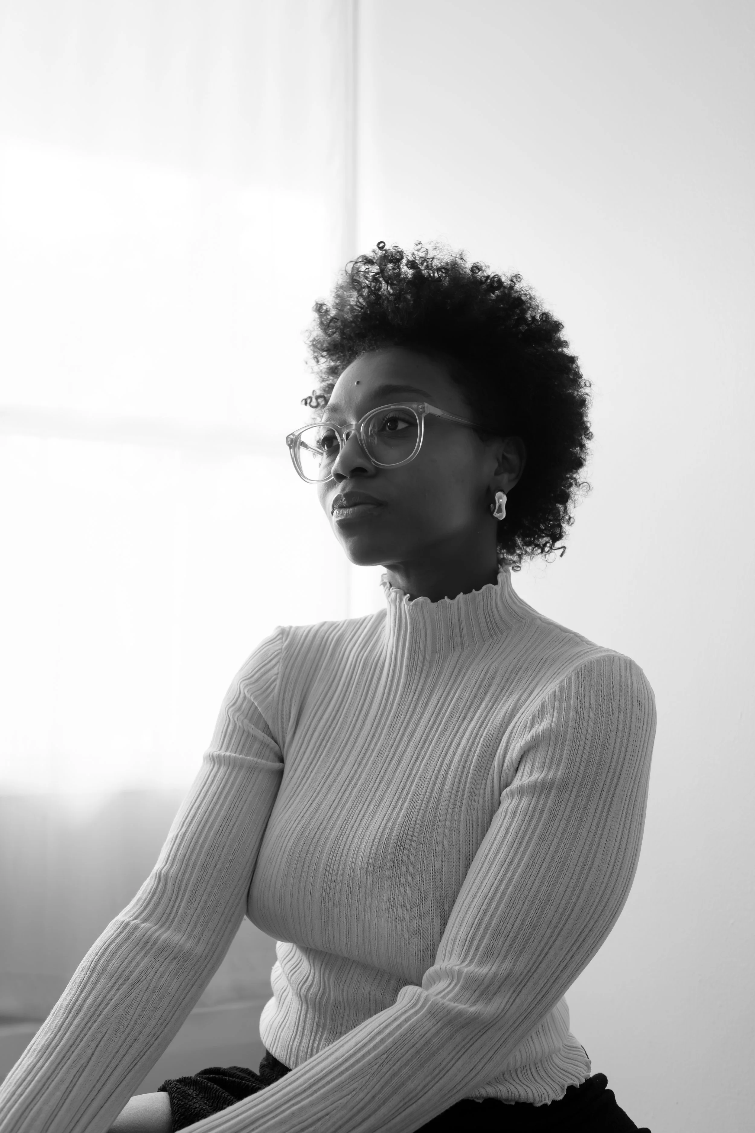 A black-and-white photo of a woman with curly hair, glasses, and earrings, seated indoors near a window, looking thoughtfully to the side.