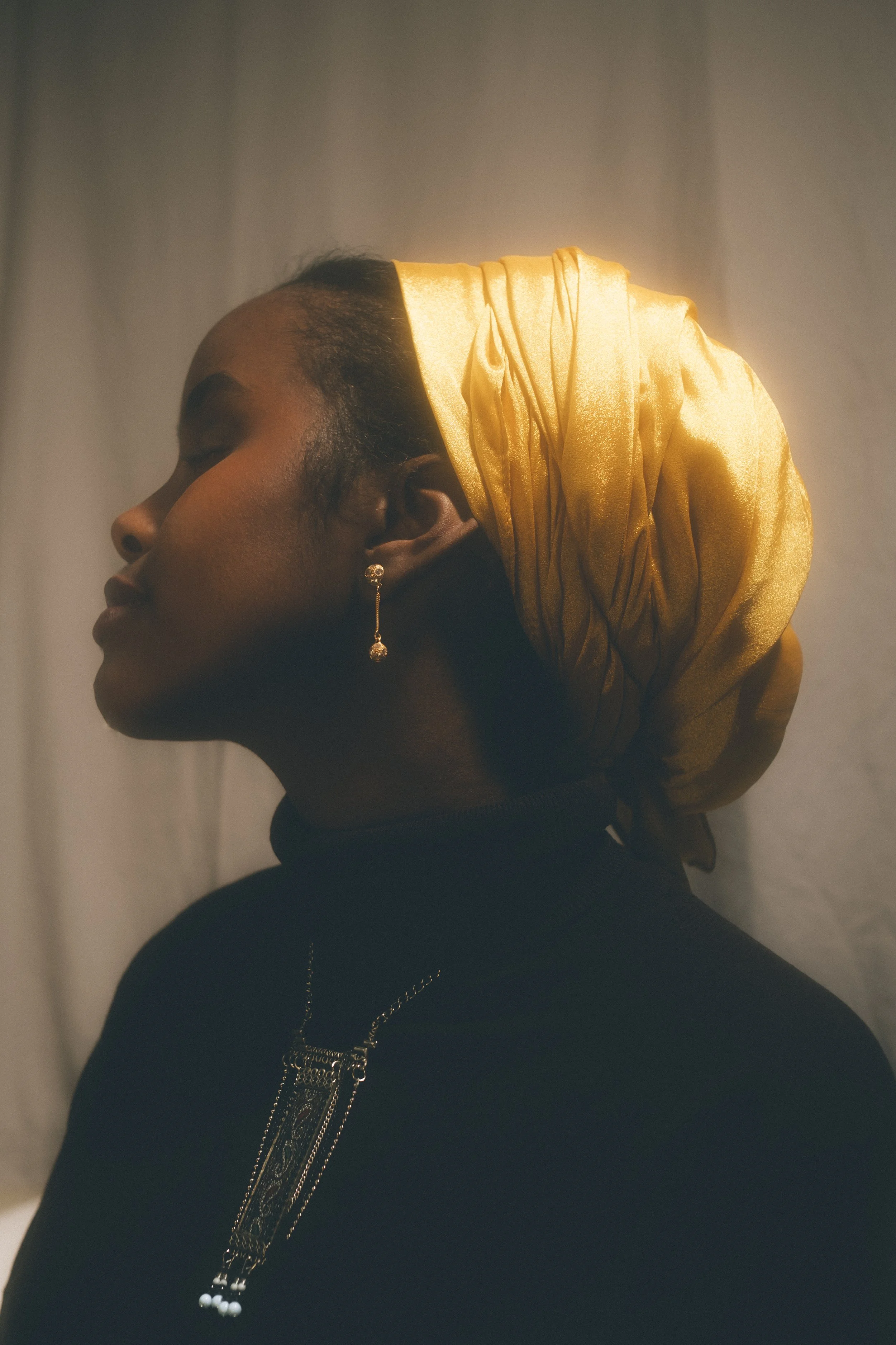 Side profile of a woman wearing a black top, gold earrings, and a large gold headwrap.