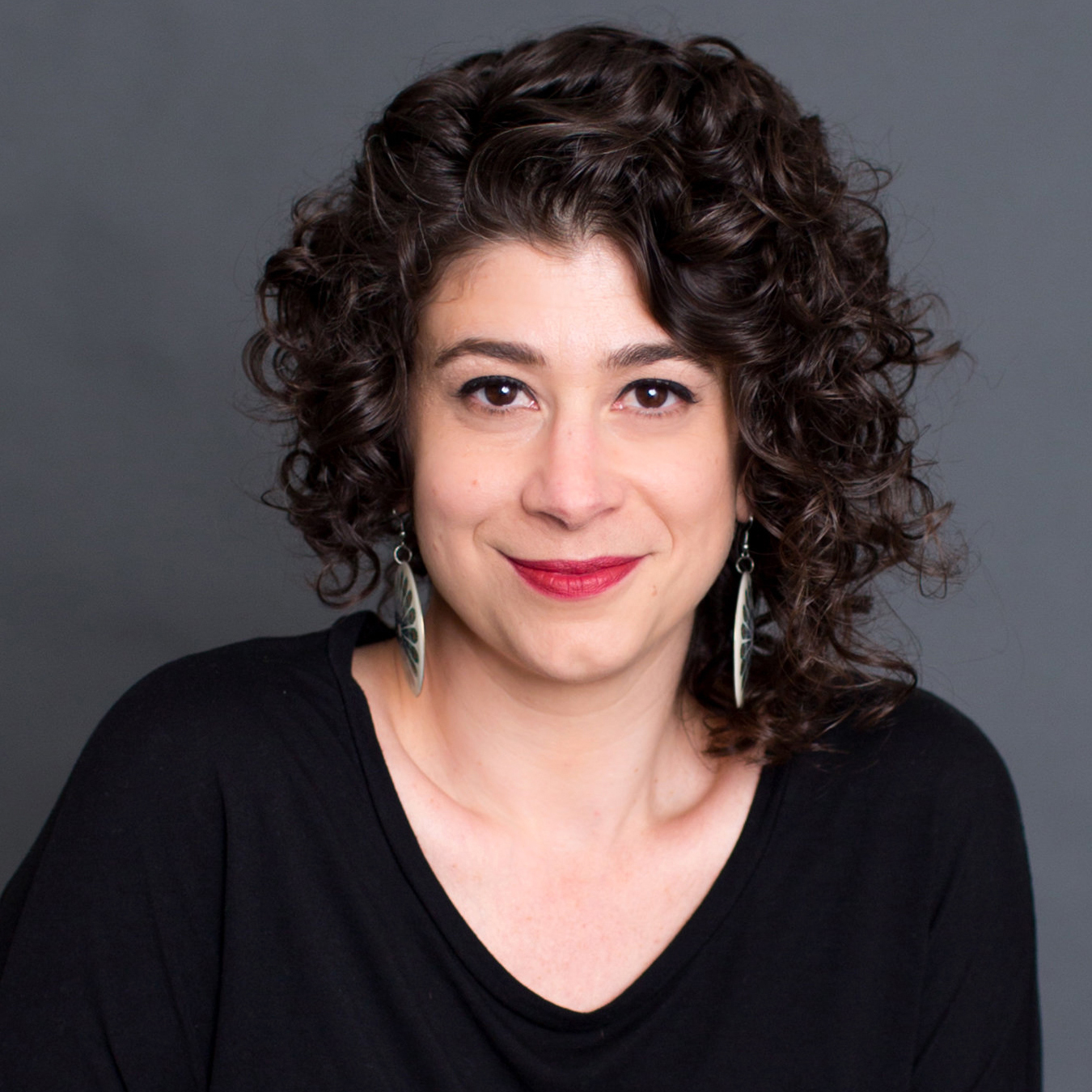 Photo of Shaleece Haas. She is pictured from shoulders up, has dark curly hair, and brown eyes. She is wearing earrings, red lipstick, and smiles at the camera.