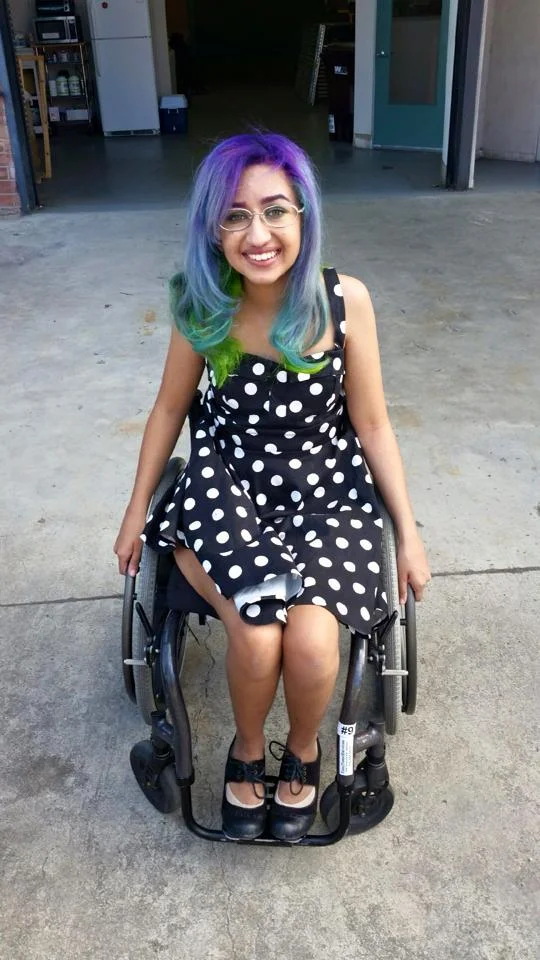 Photo of Roya Bhargava,. She is a young person of color sitting in a wheelchair. She has purple and green hair, is wearing glasses and a polka dot dress, and is smiling at the camera.