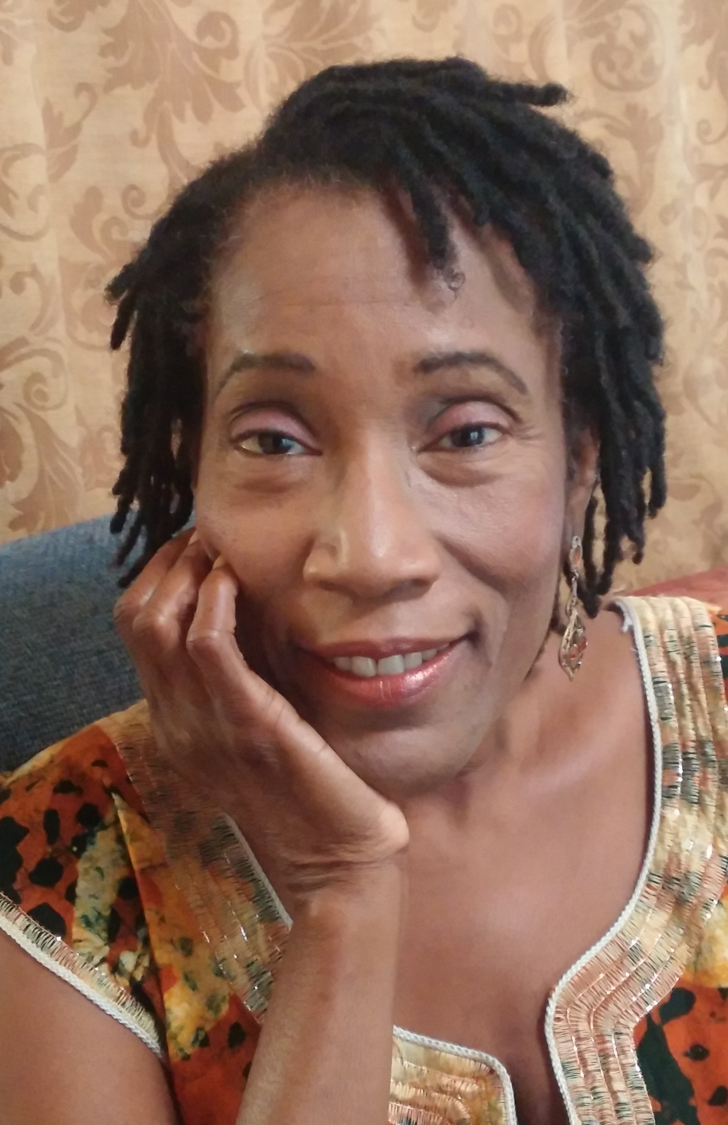 Photo of Muitma Imanii. She is an african American woman with hair in short Locks. She is wearing earrings, a brown shirt, and smiles as she leans on one hand.