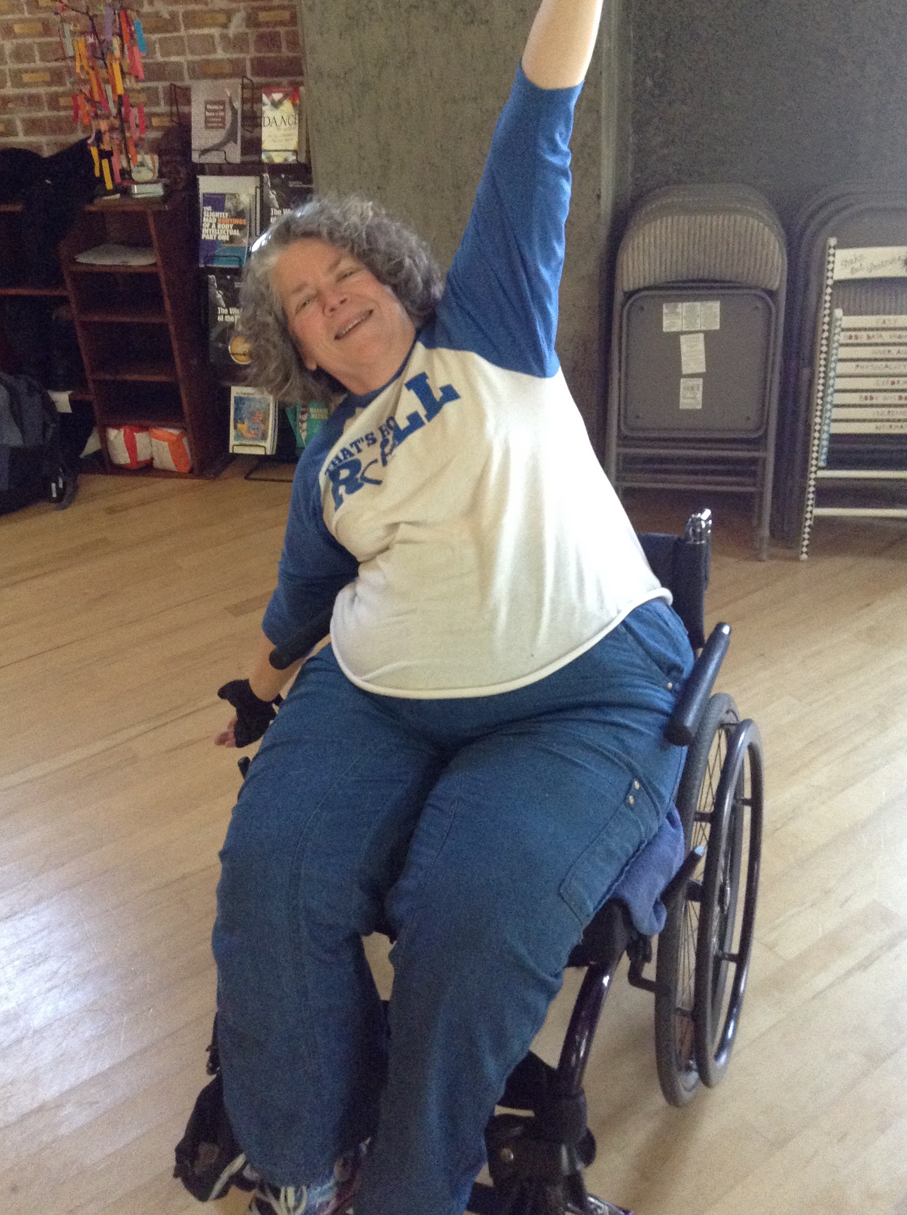 Photo of Tara Ayres. She is a white wheelchair user with graying curly hair. she sits in her wheelchair, stretching her arms out, and smiling at the camera. she is wearing a white t shirt and blue pants.