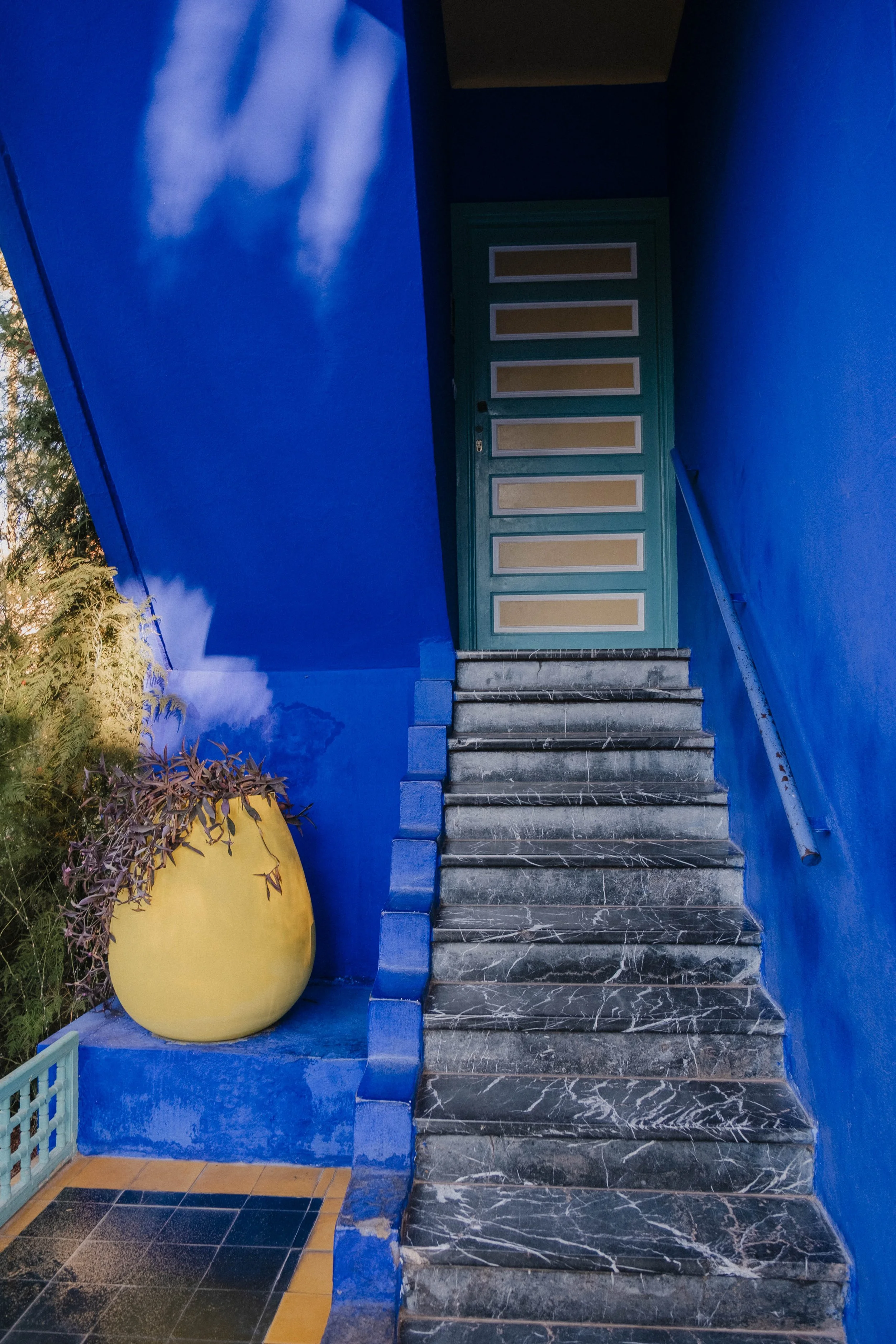 Bright blue exterior wall and staircase leading to a turquoise door with beige panels, yellow vase with purple plant on a small blue platform, and a patterned tile floor in the foreground.