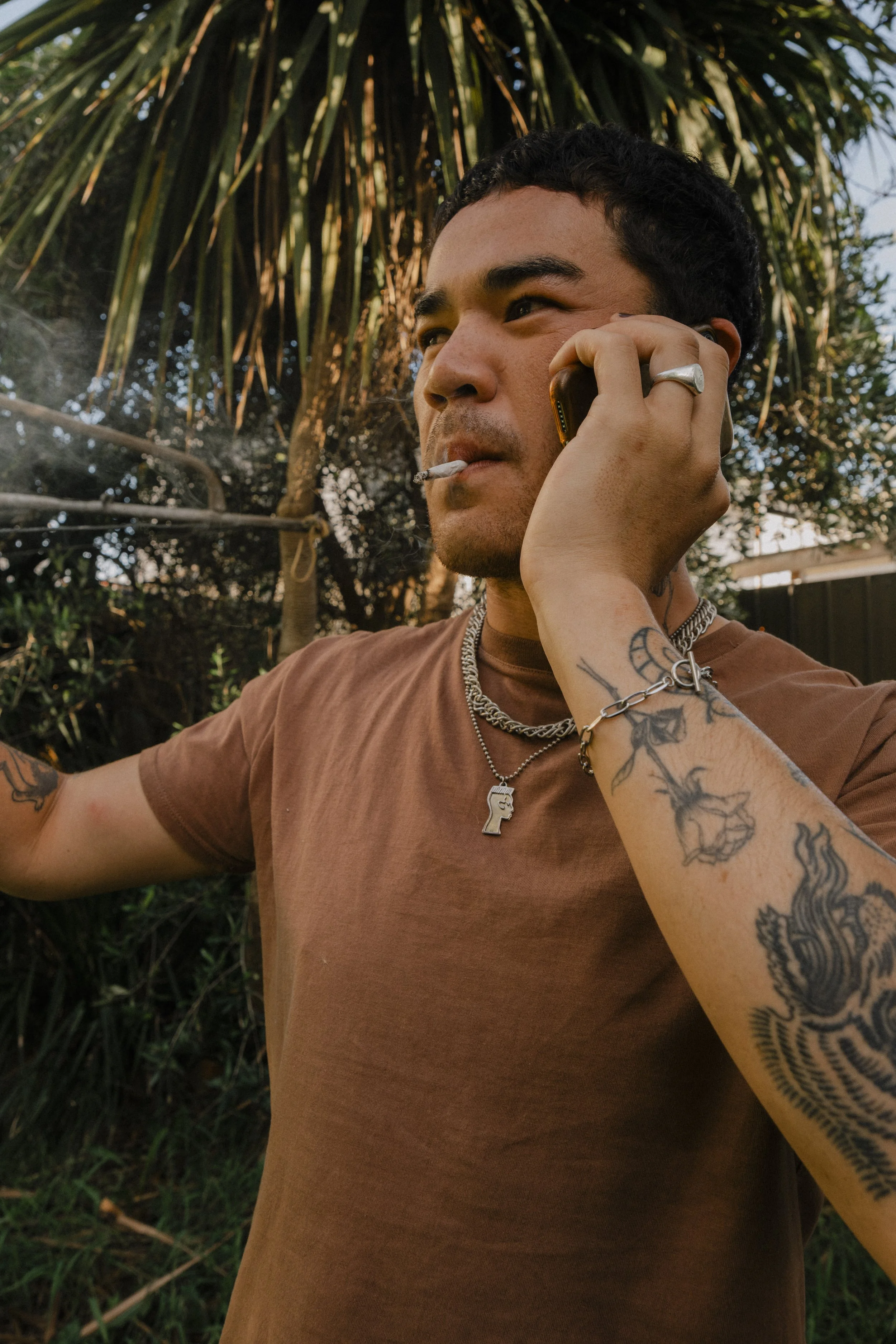 A young man with tattoos on his arm, wearing a brown T-shirt, silver chains, and a ring, is talking on a mobile phone while smoking a cigarette outdoors near some trees.