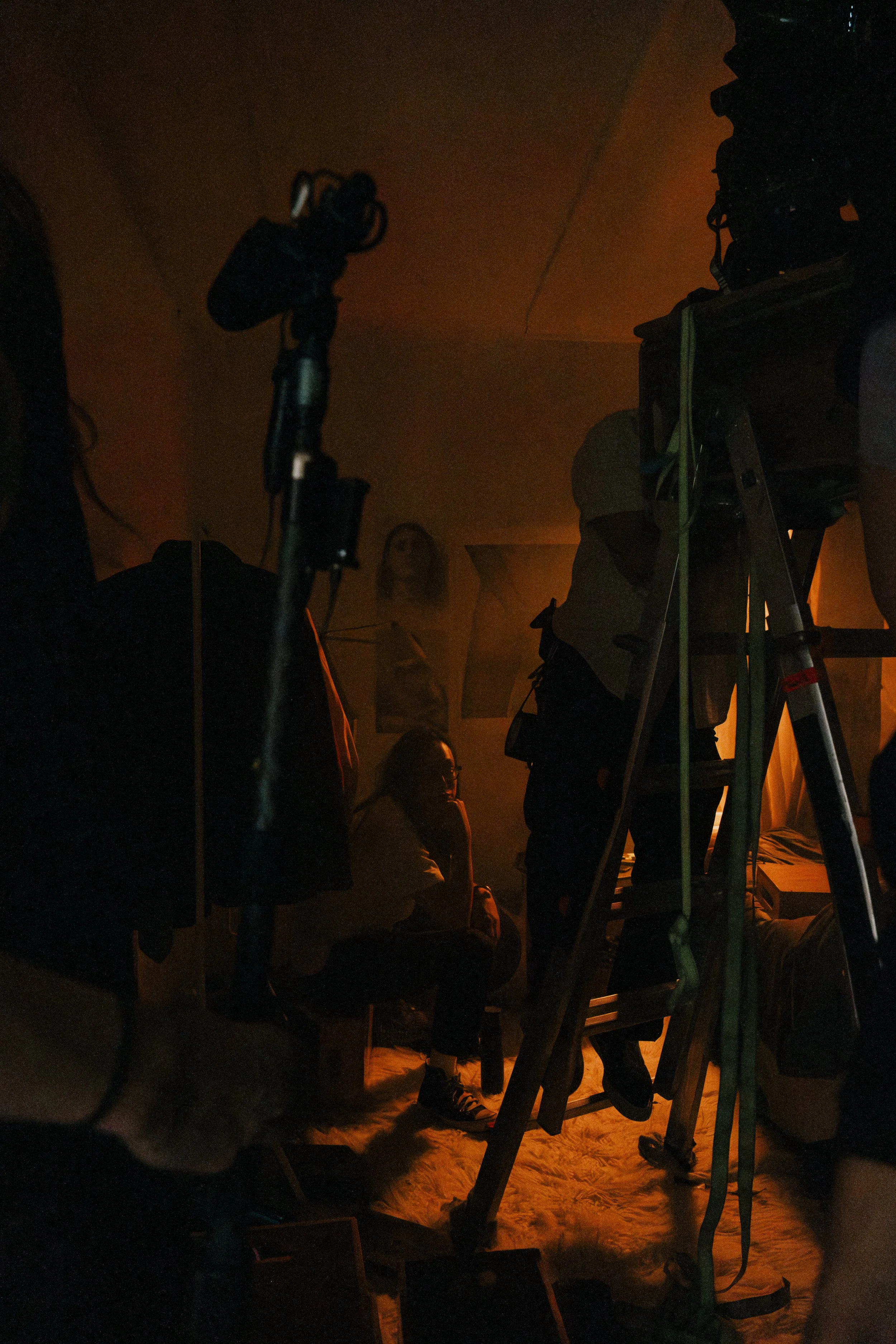 Scene with DOP sitting on a chair, surrounded by filming or photography equipment, with a woman standing in the background in a dimly lit room with orange lighting. Unit still from the short film Stranger Brother.