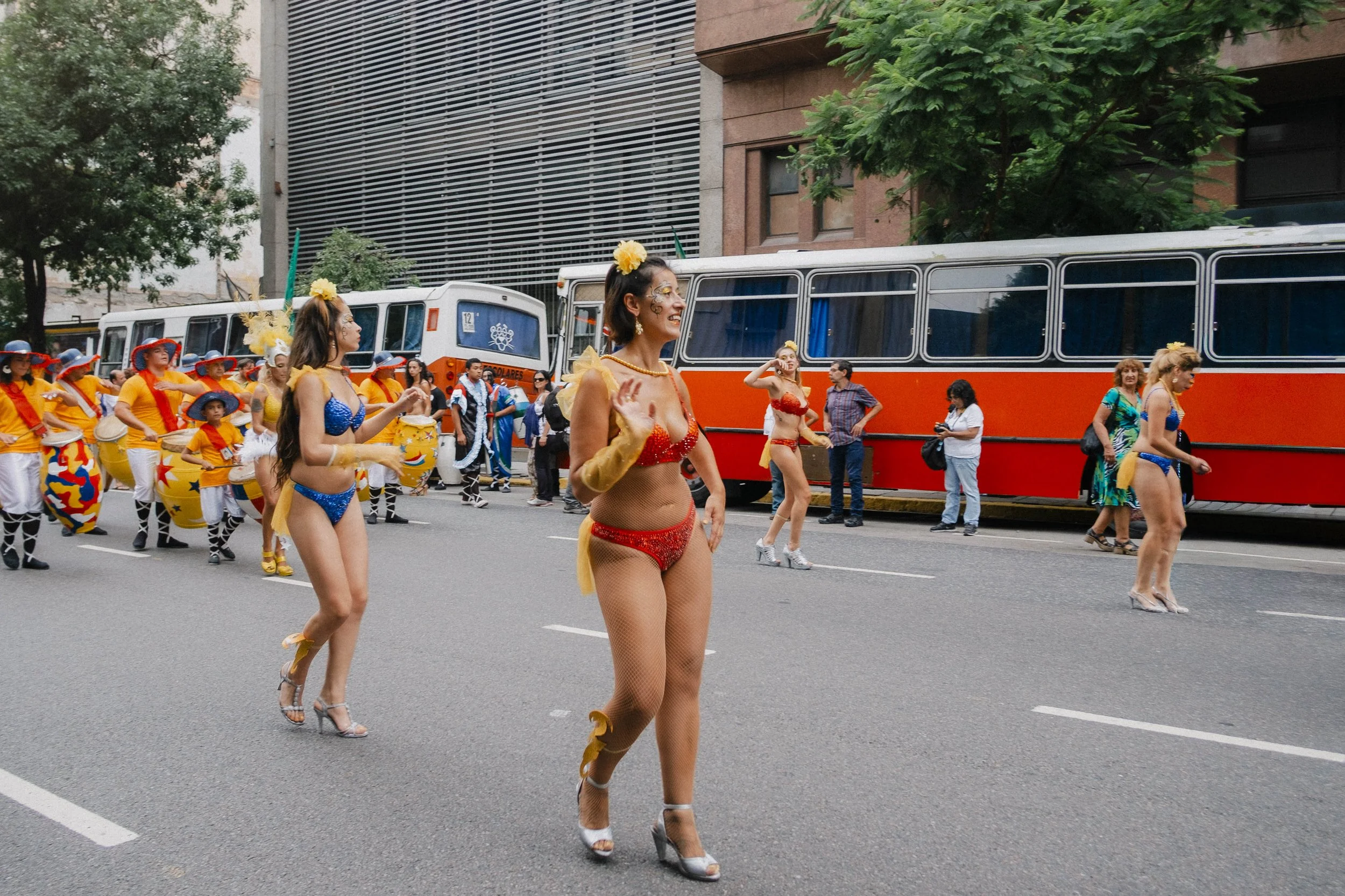 Women and children in colorful costumes participating in a parade on city street, with buses and spectators in background.