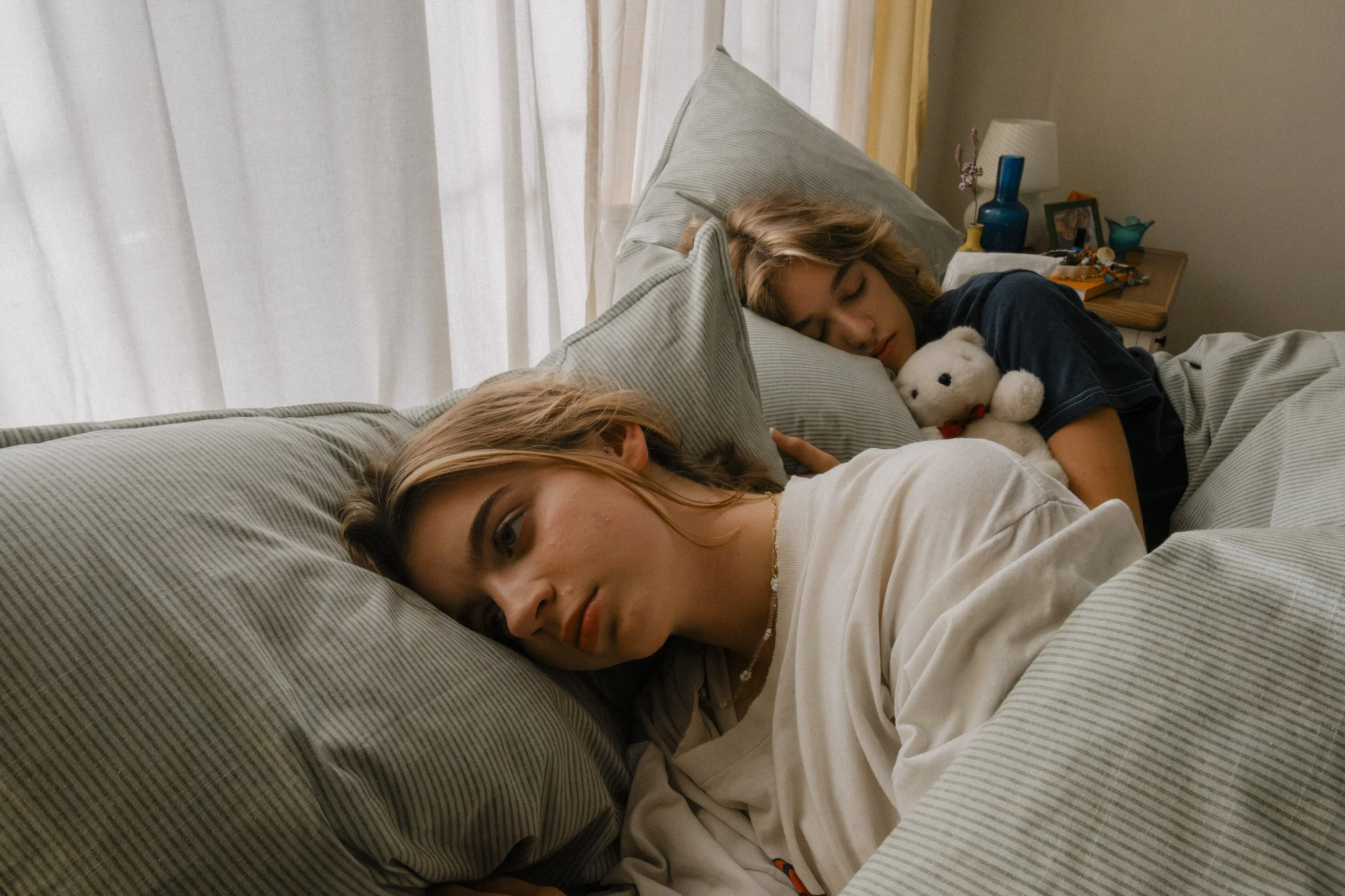 Two young women sleeping in bed, one resting her head on a pillow, holding a stuffed animal, with a cozy room in the background.