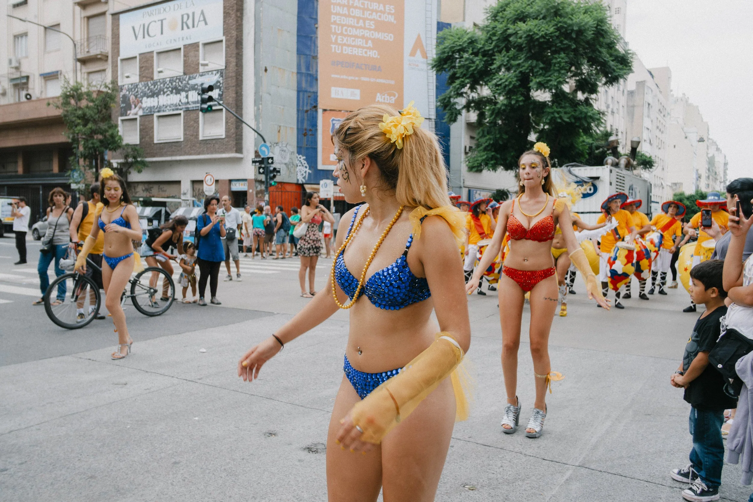 Three women in colorful bikinis performing in a street parade, with a crowd of spectators and dancers in the background.