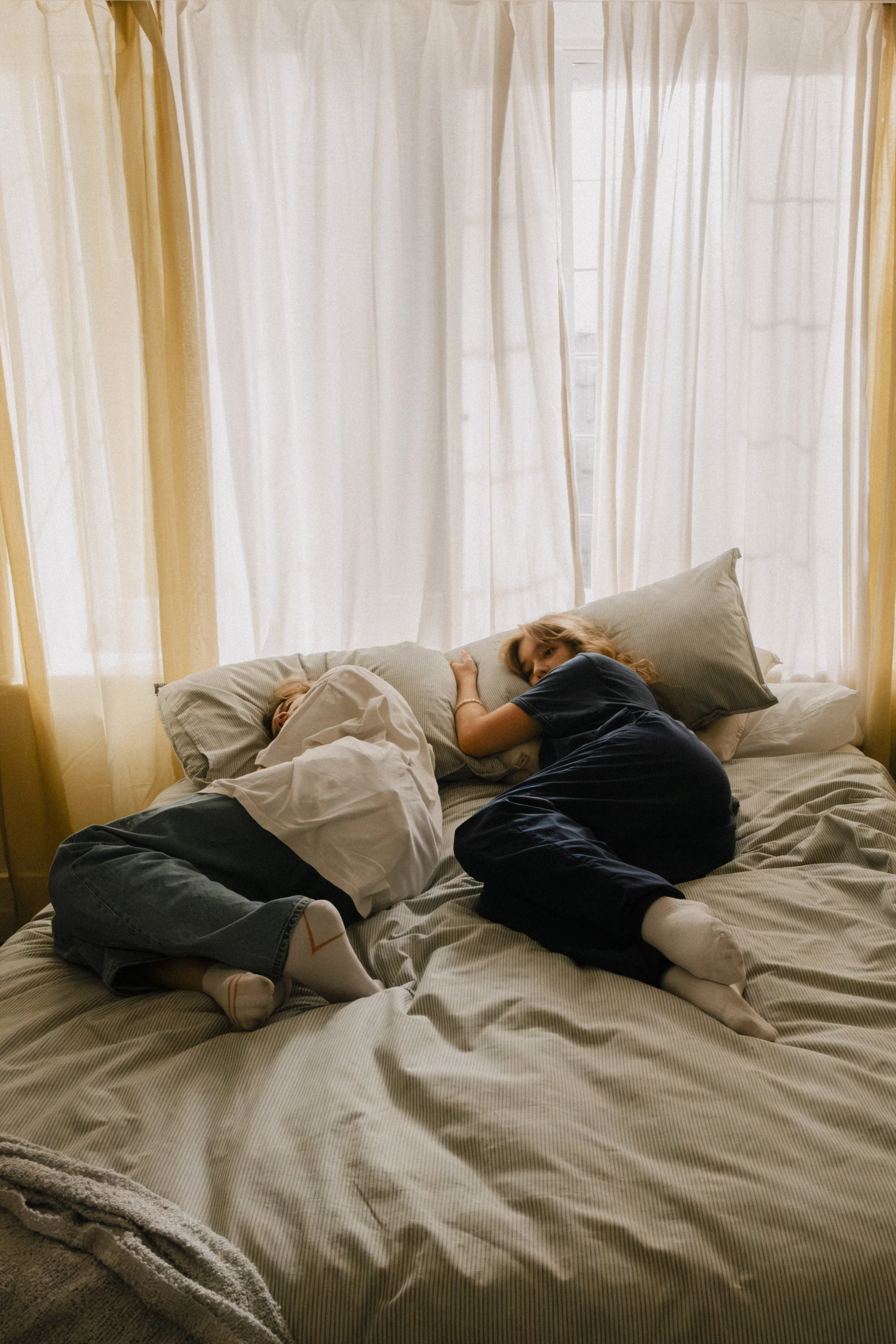 A mother and her young child sleeping together on a bed with cream-colored sheets and beige pillows, near large windows with sheer white curtains.