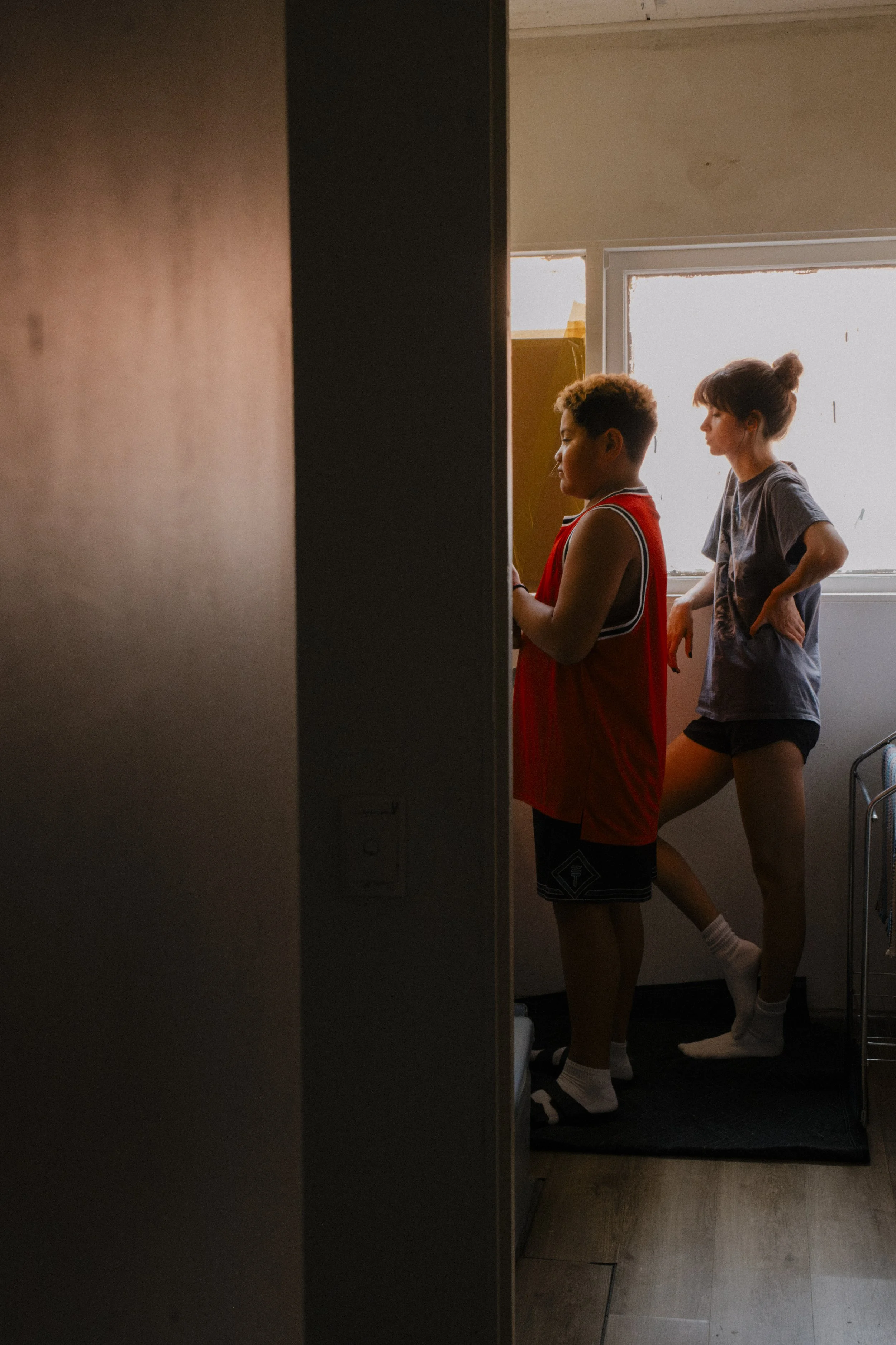 Two peop stand near a window in a dimly lit room, one wearing a red sleeveless jersey and the other in a gray T-shirt and black shorts, both looking toward the window, unit still from the short film Stranger Brother.