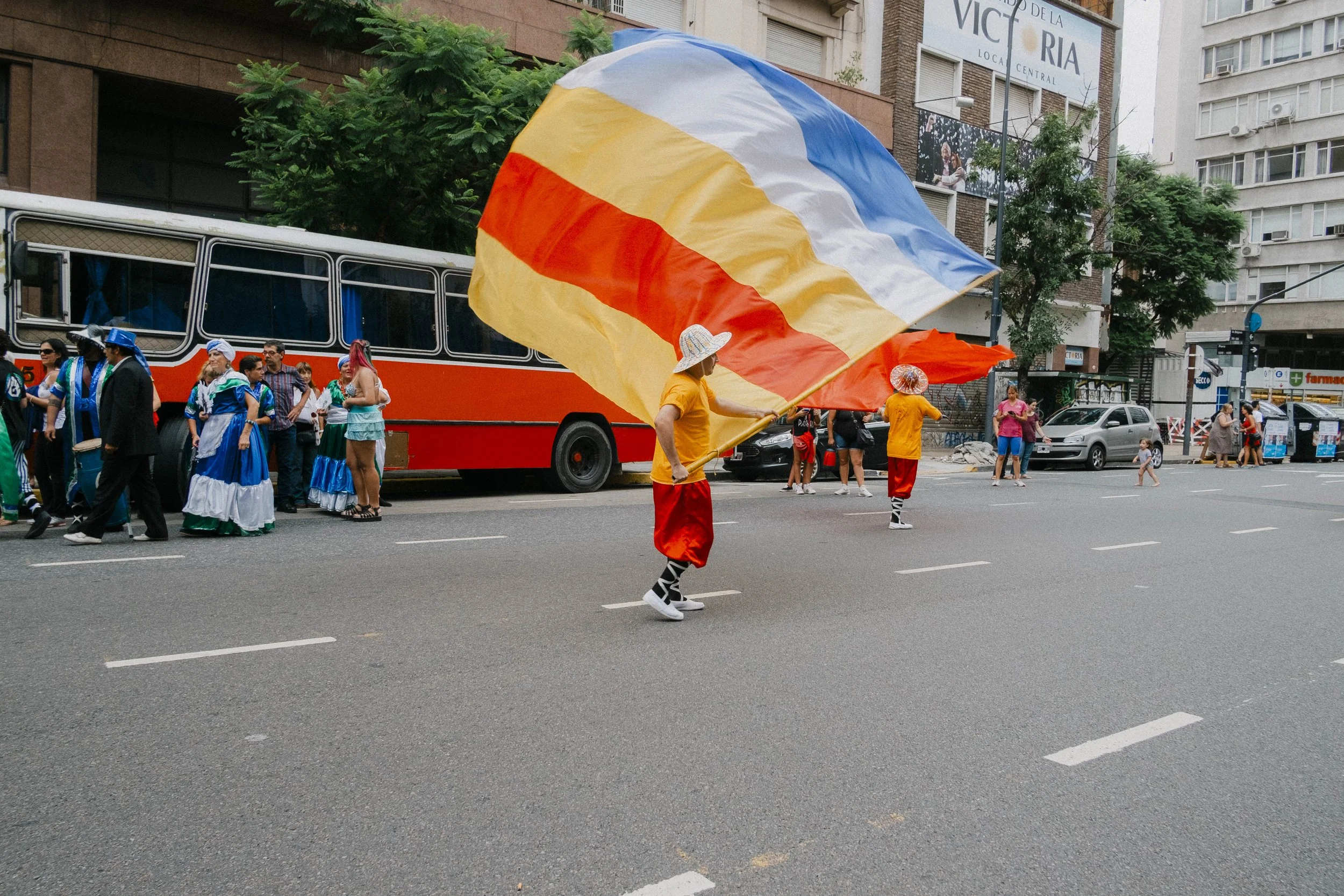 People participating in a parade, some dressed in traditional costumes, with one person waving a large tricolor flag on a city street.