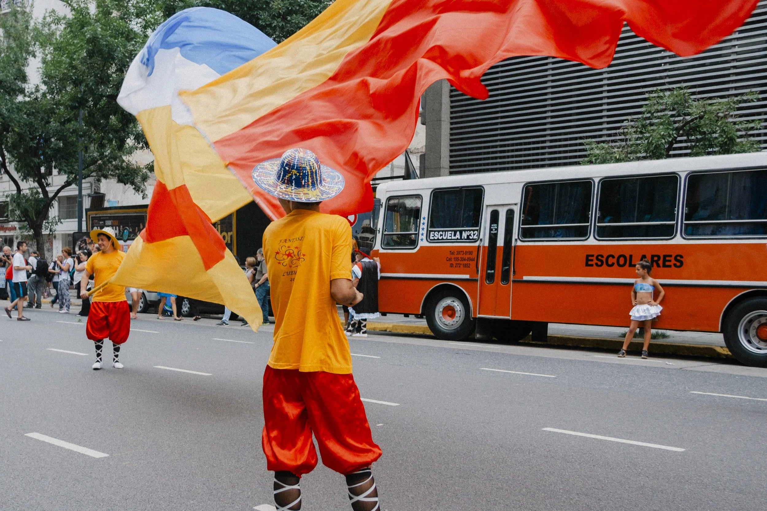 A parade scene with performers in bright yellow and red costumes, one with a sombrero, waving large yellow and orange flags. A girl in a blue and white outfit stands near an orange bus labeled 'Escuela N° 32'. There are spectators lining the street and trees in the background.