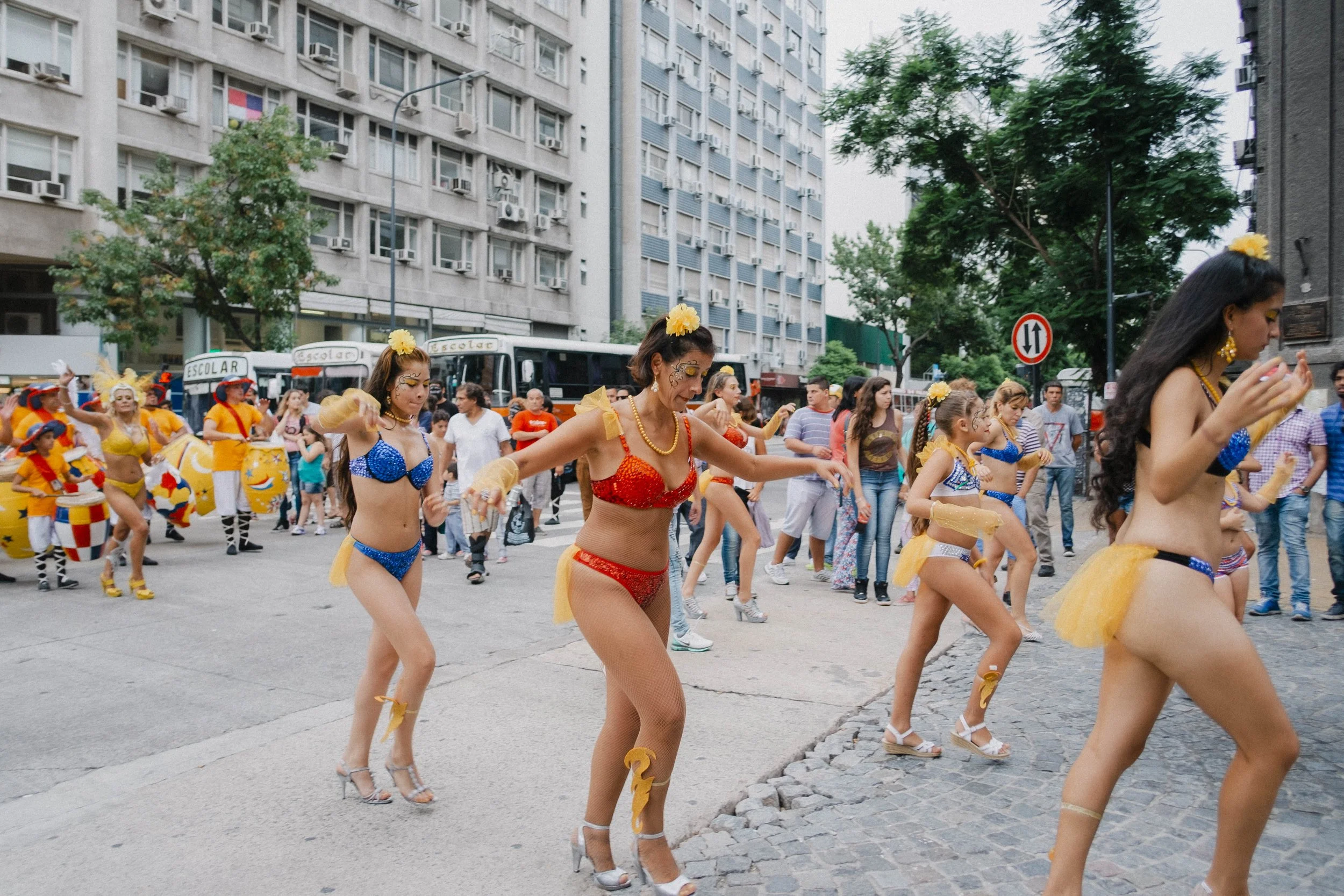 Women dressed in colorful bras, tutus, and high heels dance in a street parade, with onlookers and city buildings in the background.