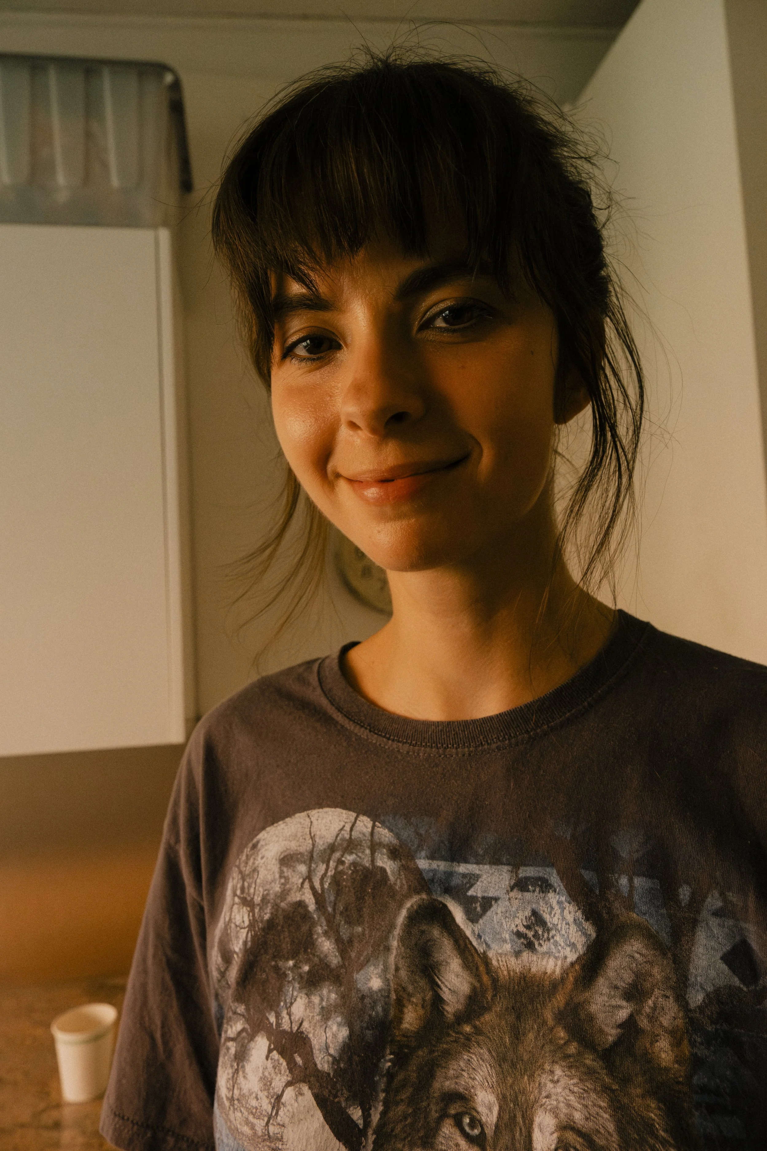 A woman with short dark hair and a slight smile, wearing a graphic t-shirt with a wolf design, standing in a room with white cabinets and a countertop.
