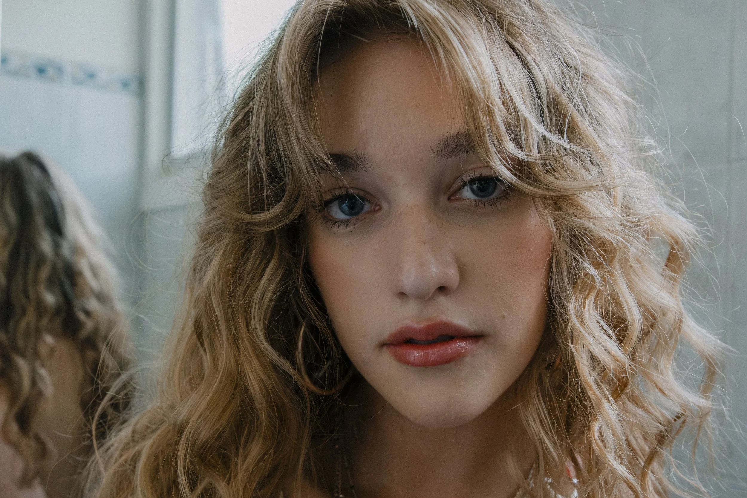 Close-up of a young woman with curly blonde hair and blue eyes, looking directly at the camera.