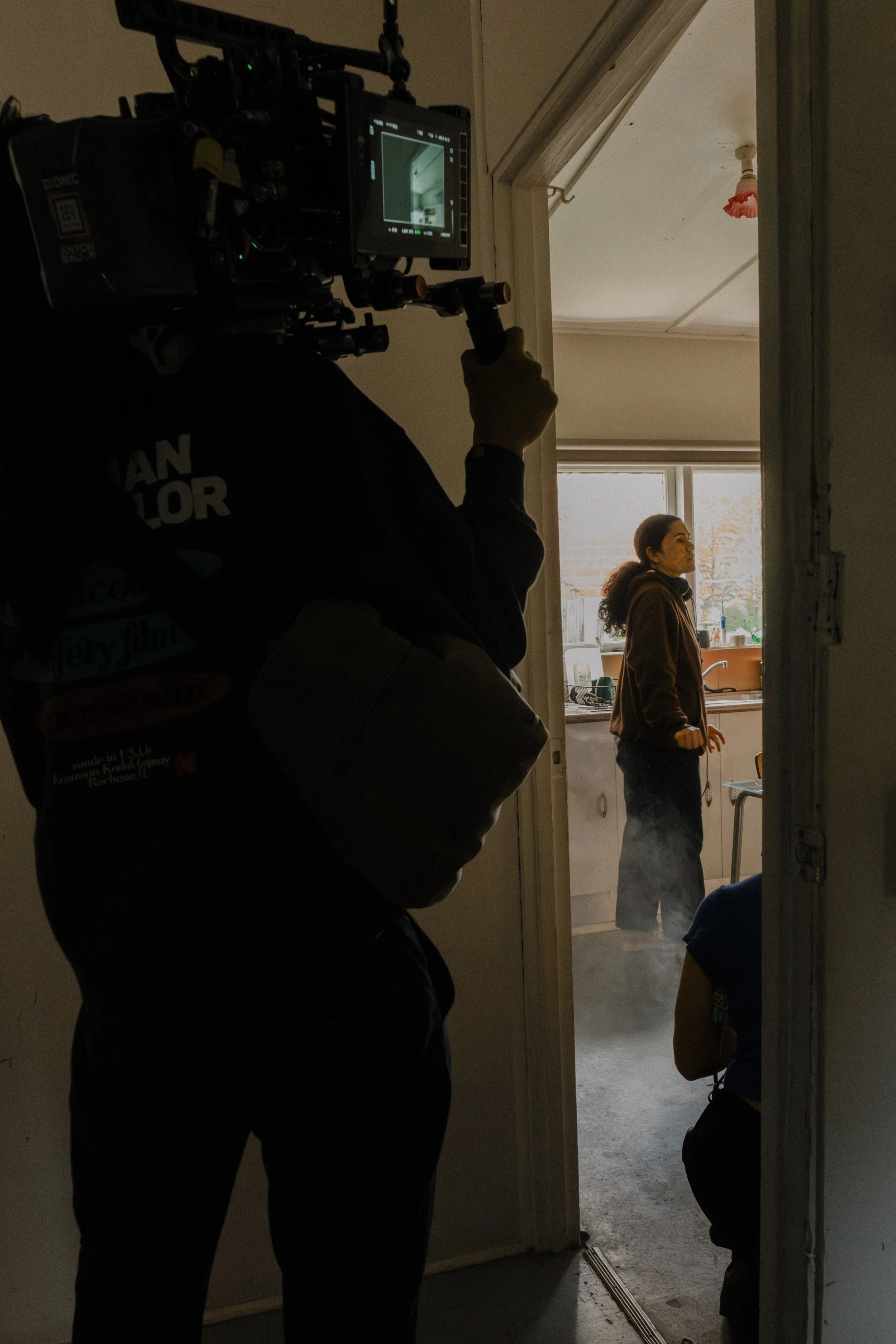 DOP operating a camera with a large lens, capturing a woman standing in a kitchen near a window, with another person partially visible kneeling nearby. Unit still from the short film Stranger Brother.