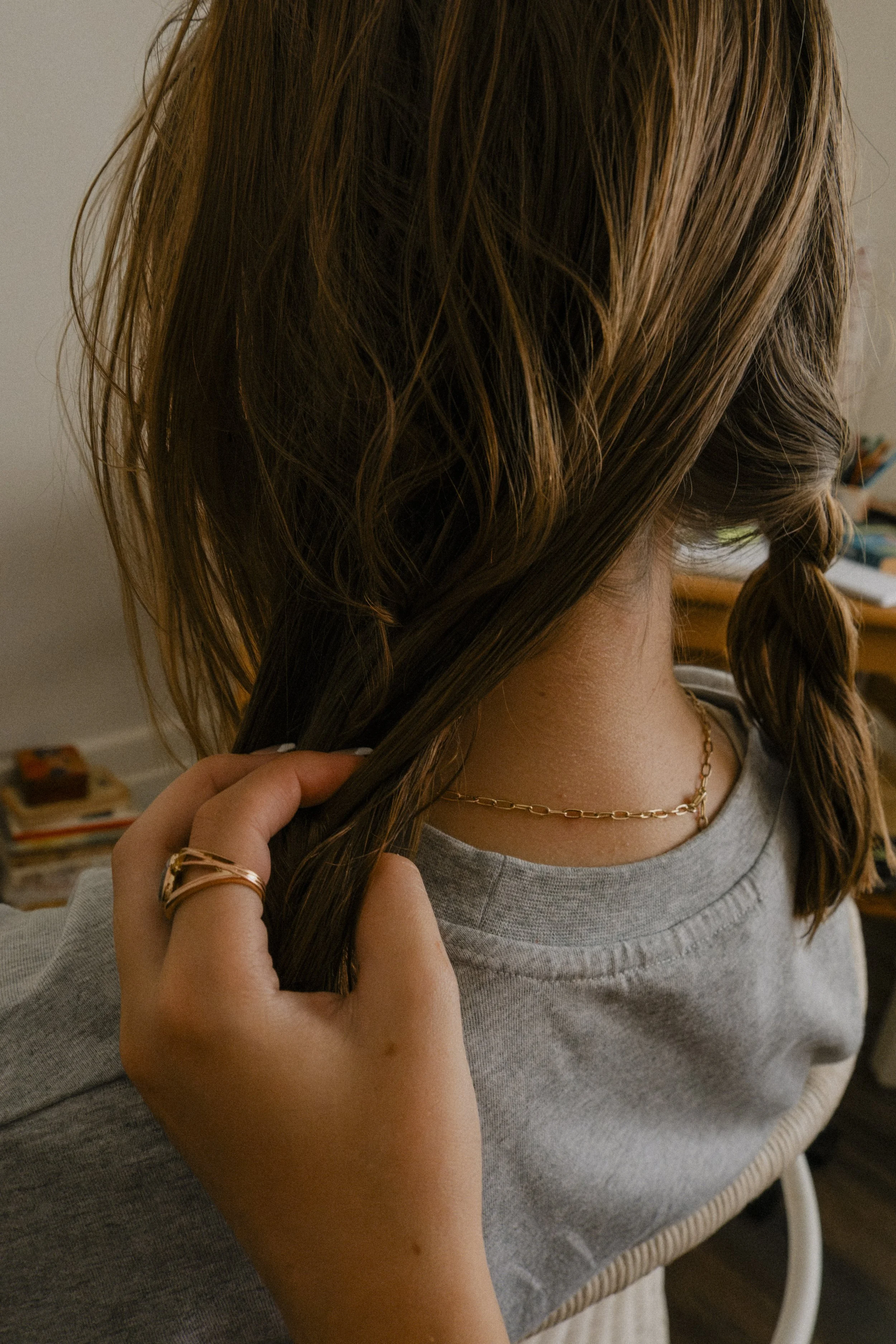 A person with brown, wavy hair wearing a gray sweatshirt, gold necklace, and rings, holding strands of their hair.