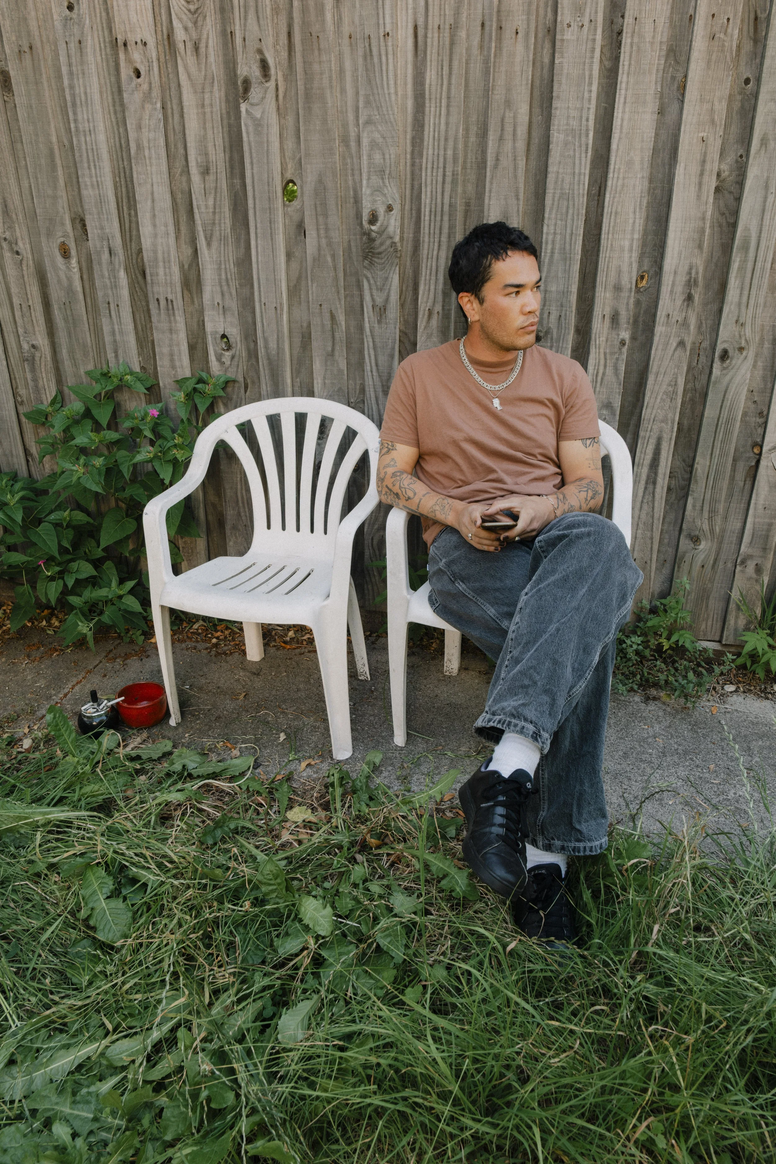 A young man with tattoos, wearing a brown T-shirt, black jeans, and black sneakers, sitting on a white plastic chair outdoors, holding a phone, with an empty matching chair next to him, plants, and a wooden fence behind him.