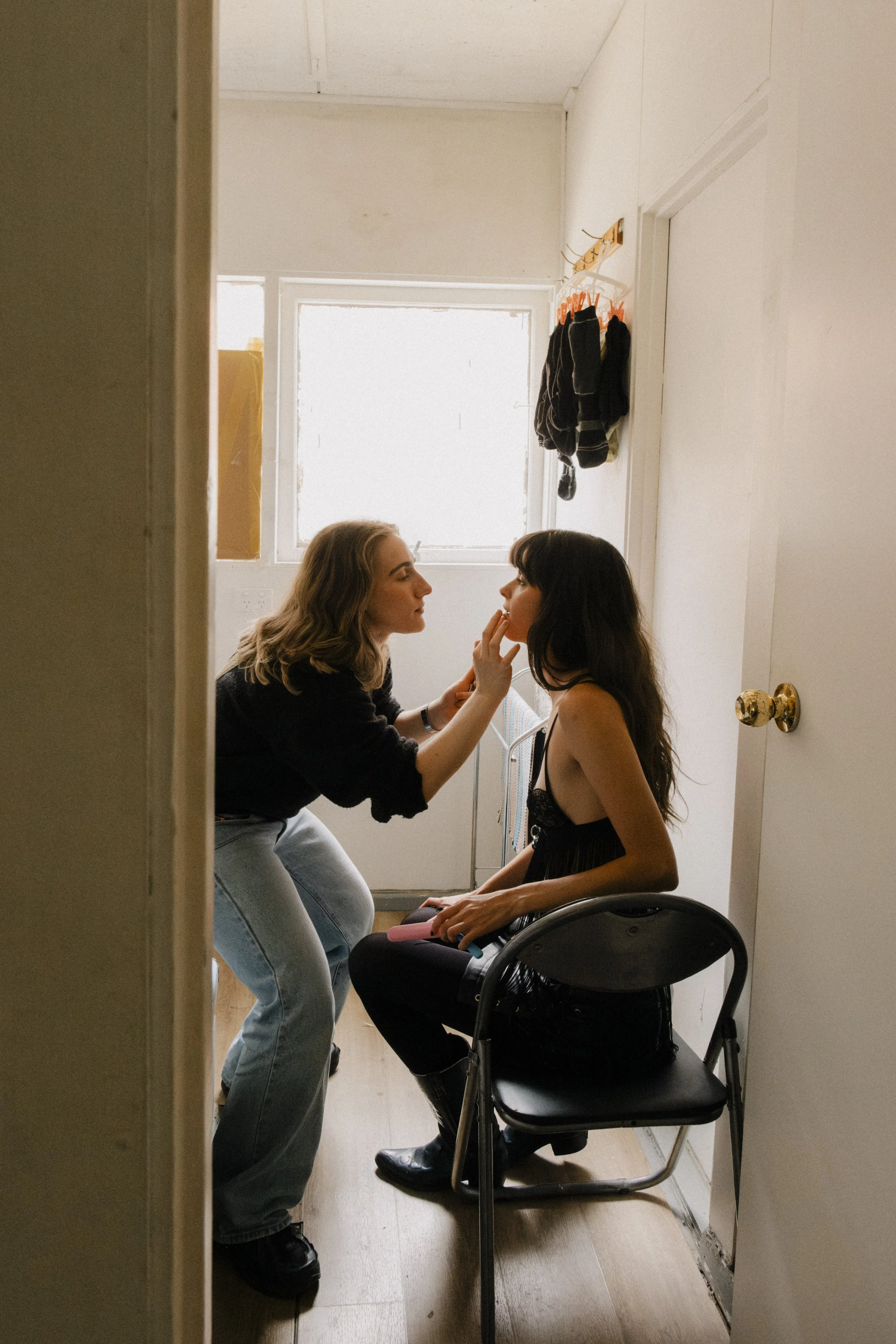 Two women are in a small, brightly lit room. One woman, sitting on a chair, is getting her makeup done by the other woman, who is standing and carefully applying makeup to her face. Unit still from the short film Stranger Brother.