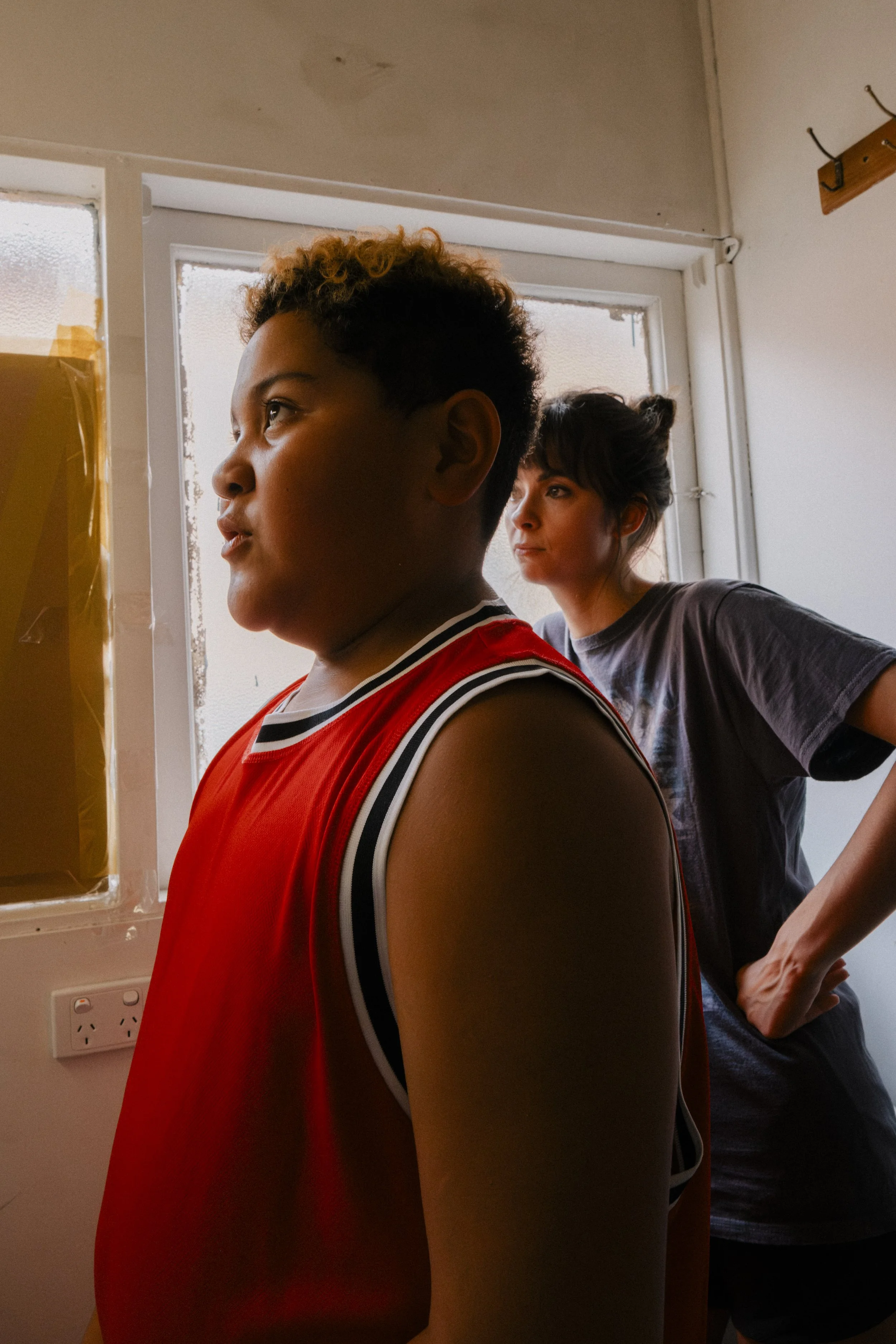 A young boy in a red sports jersey standing indoors near a window, with a woman in a gray t-shirt standing behind him looking out the window. , unit still from the short film Stranger Brother.