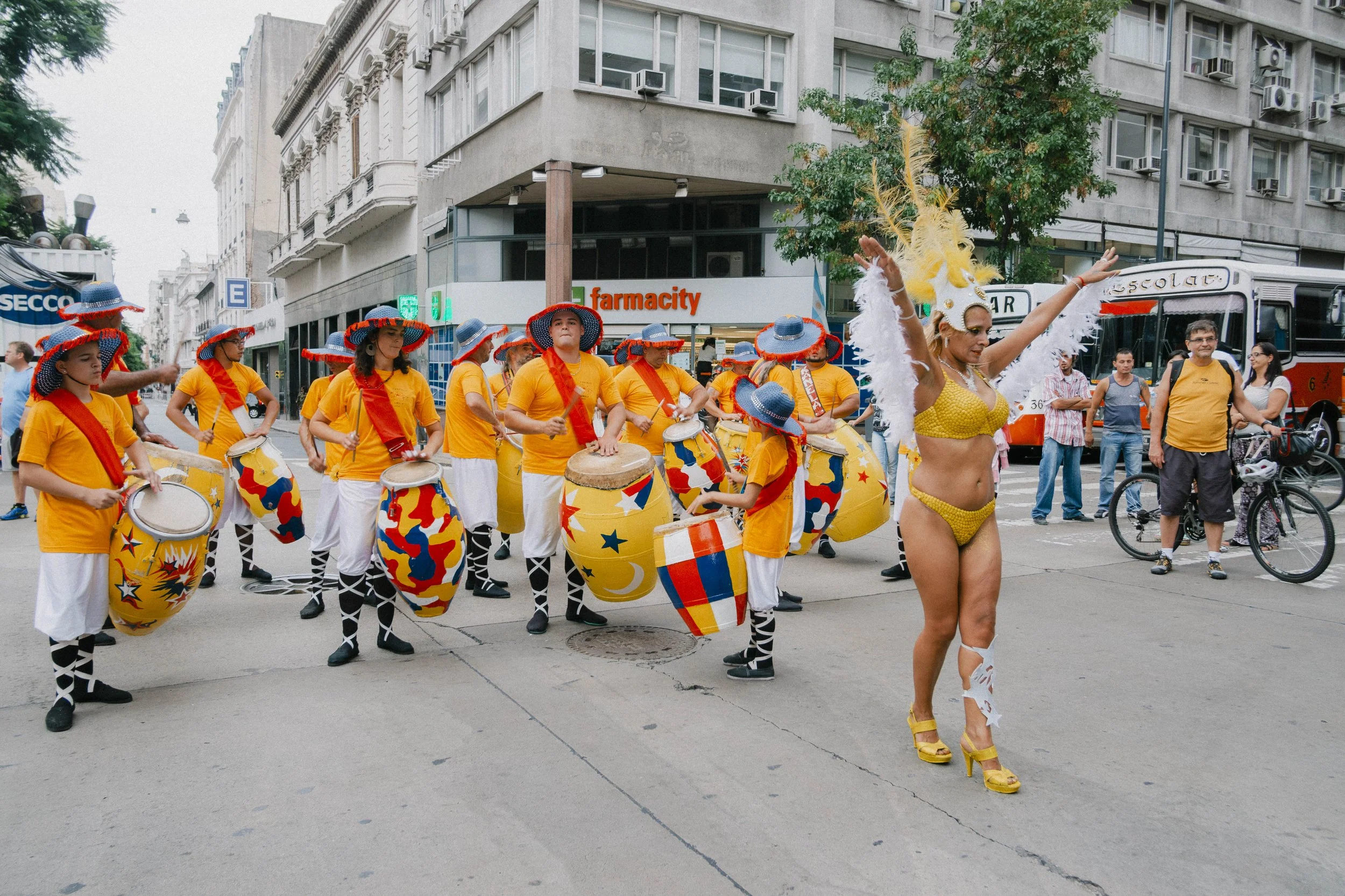 A street parade featuring performers in colorful costumes. A group of drummers in yellow shirts, white pants, and wide-brimmed hats decorated with red, blue, and yellow, are playing drums. A dancer in a yellow swimsuit, high heels, and a feathered headdress is dancing with her arms raised. Spectators are watching, and city buildings, including a pharmacy, and a bus are visible in the background.