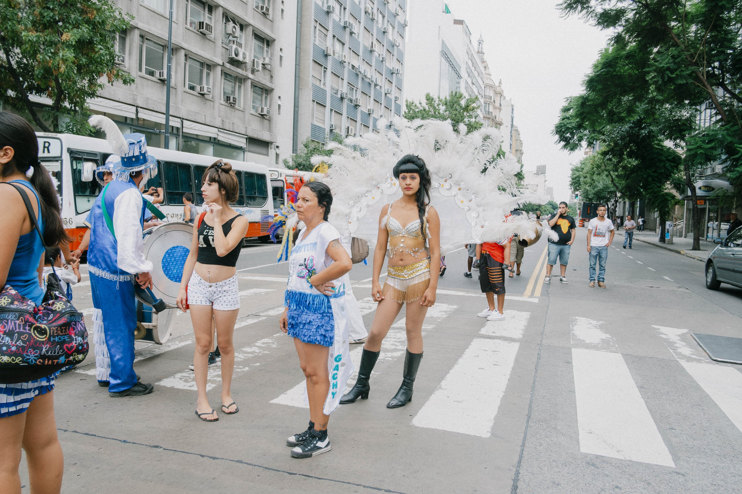 People walking and standing on a city street during a parade or festival, including a woman in an elaborate feathered costume with black boots, and a man in a colorful costume with a large hat, with buildings and trees in the background.