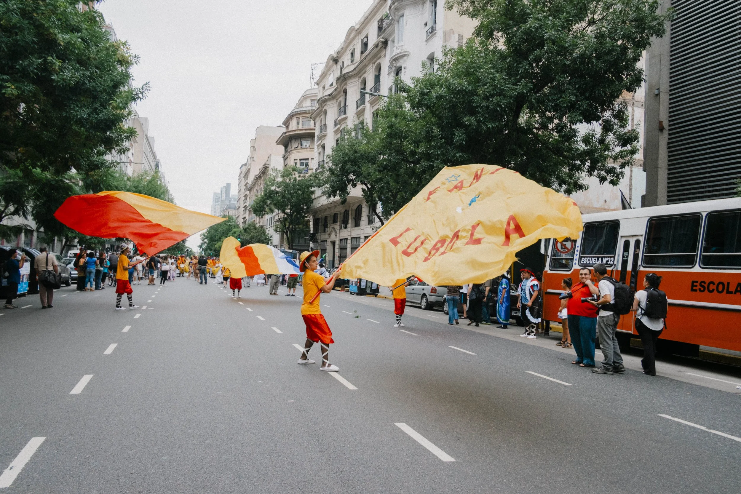 People participating in a parade on a city street, waving large yellow and red flags, with some wearing red and yellow uniforms, and others standing beside orange buses.