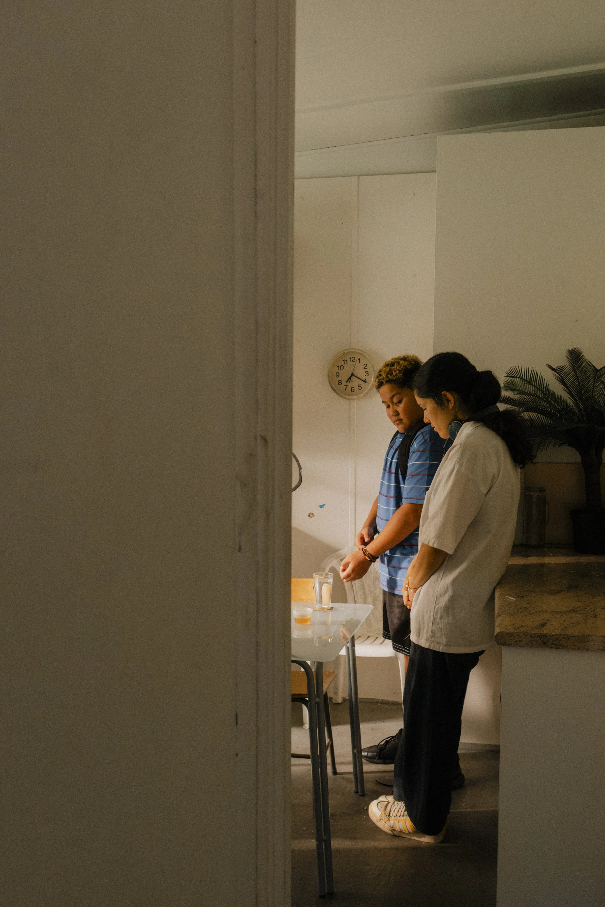 A nurse and a young man stand at a table in a kitchen, both looking down at the table with a clock on the wall behind them.