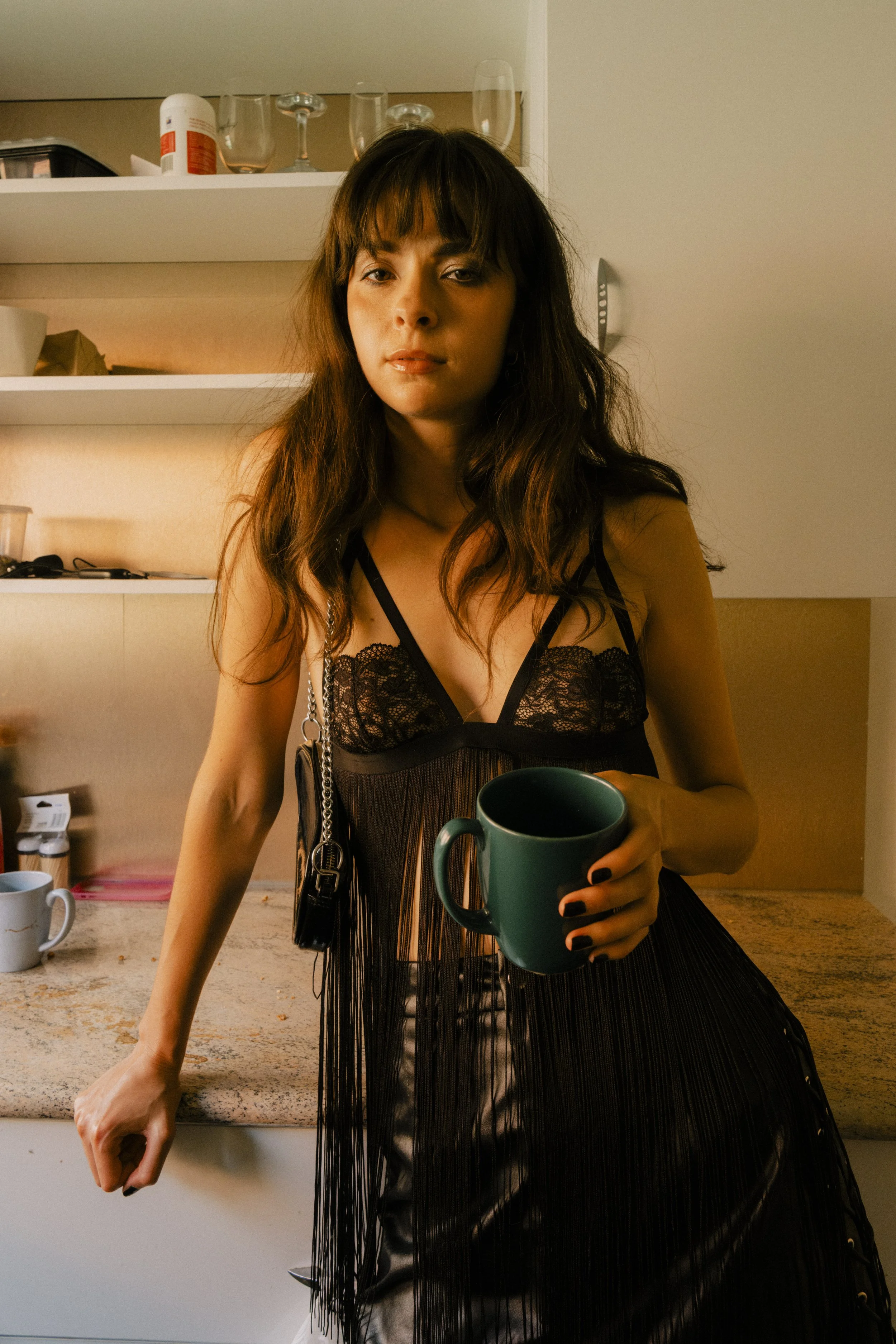 A young woman with long dark hair and bangs, wearing black lace lingerie and a black fringed dress, holding a teal mug, standing in a kitchen.