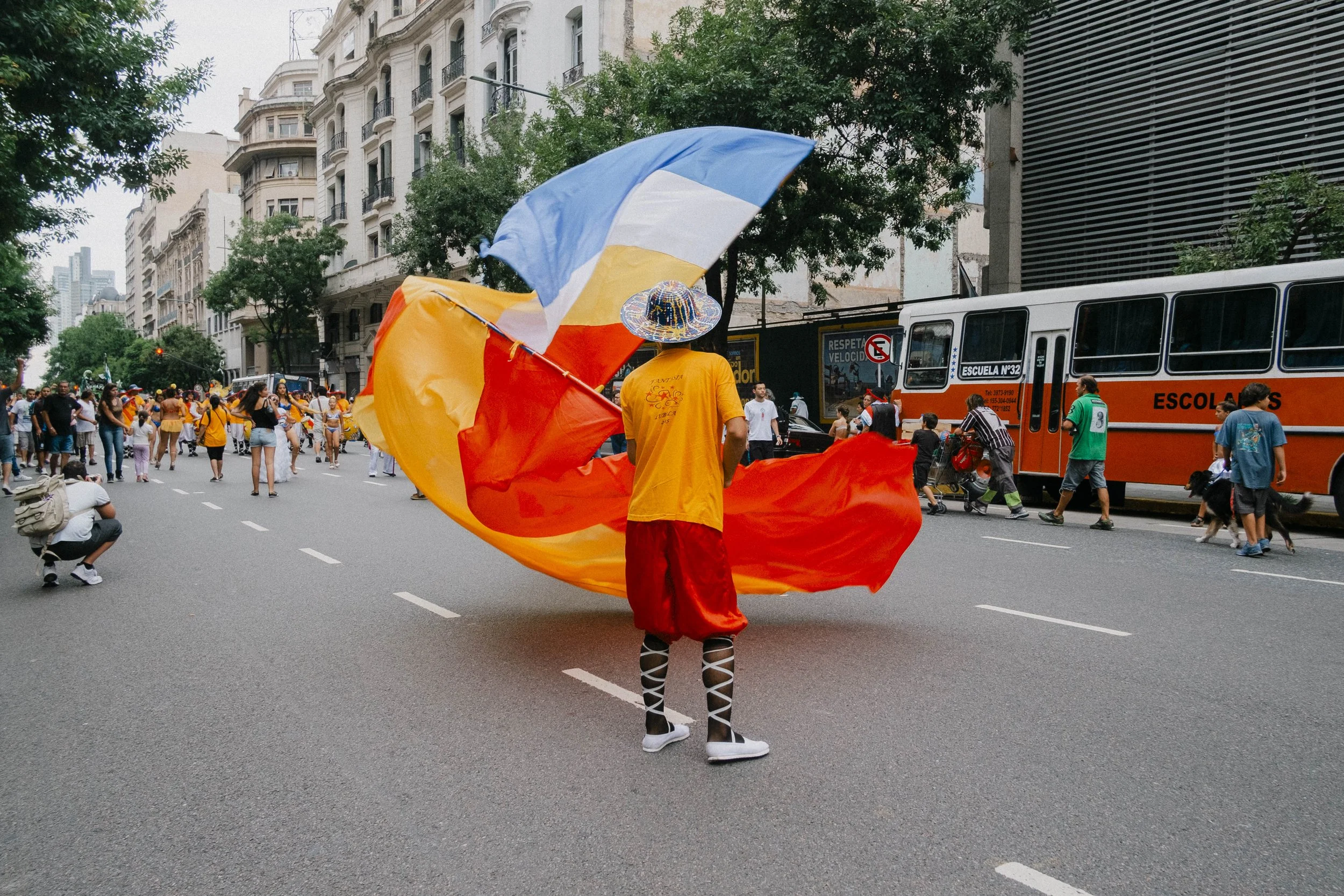 Person dressed in colorful costume with a large rainbow flag on a city street, participating in a parade or protest, with other people and a bus in the background.