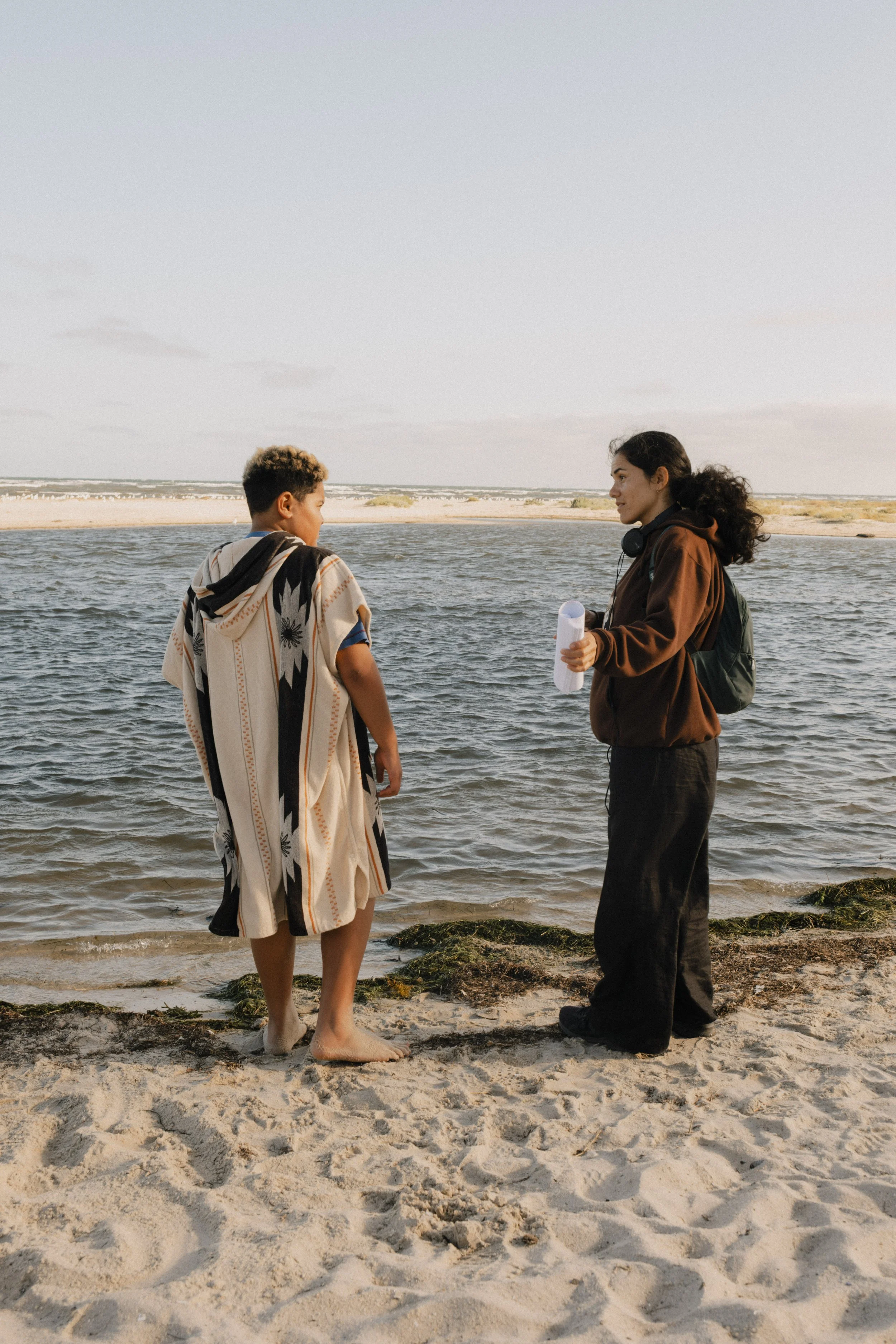 Two people are having a conversation on the beach near the water, one holding a rolled-up paper, with sandy and rocky shore and ocean in the background. Unit still from the short film Stranger Brother.