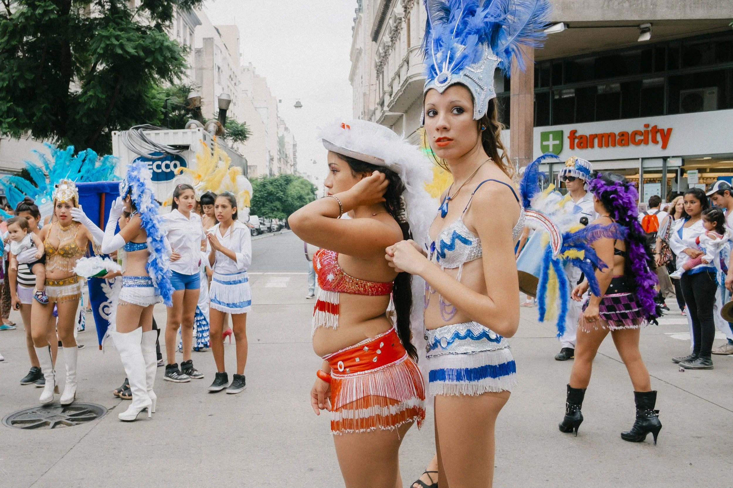 Women in colorful costumes and feathered headdresses participating in a street parade, with spectators watching nearby.
