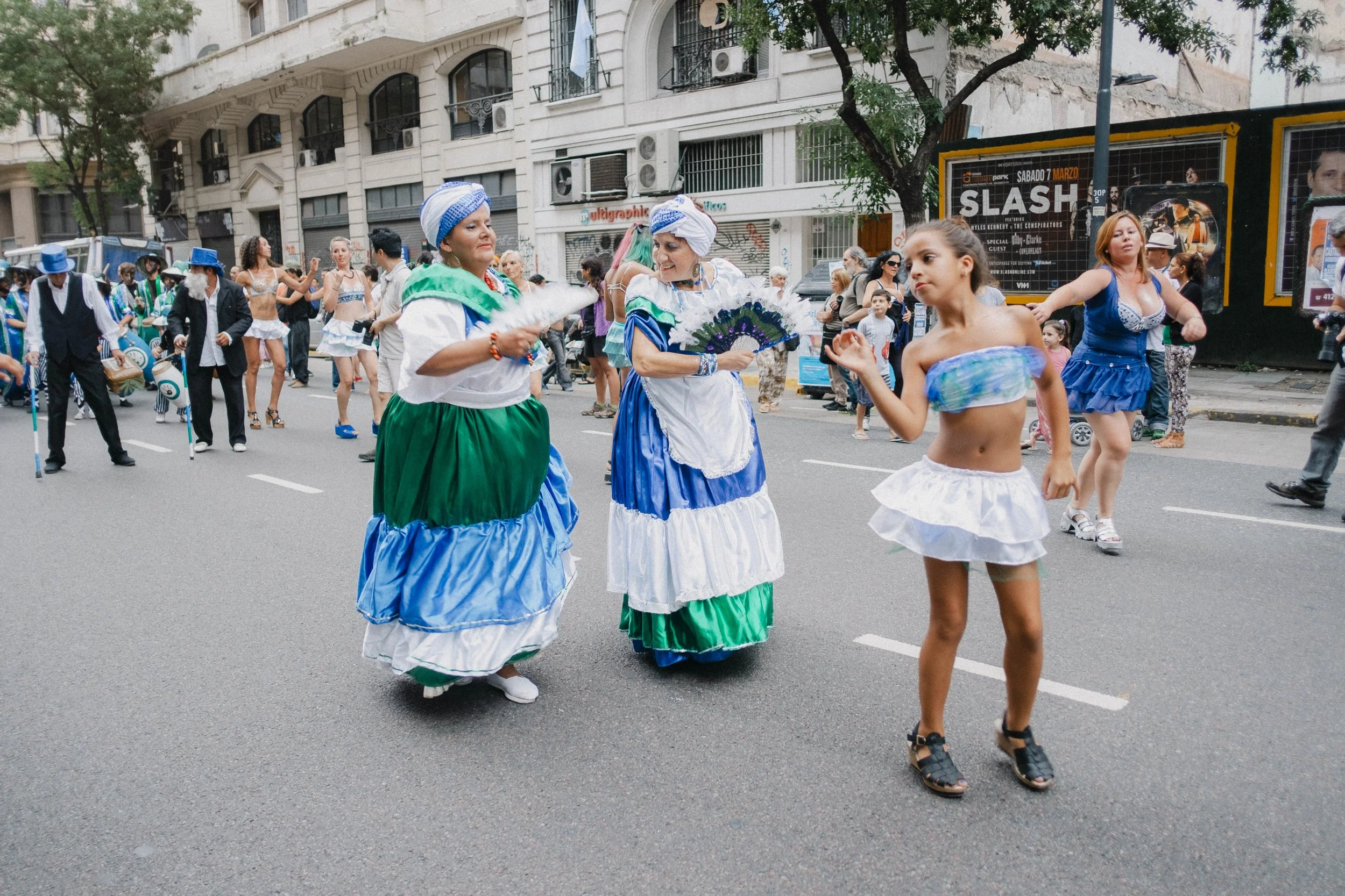 People participating in a street parade, with women dressed in traditional colorful costumes, and a girl dancing in a modern blue outfit, with onlookers and buildings in the background.