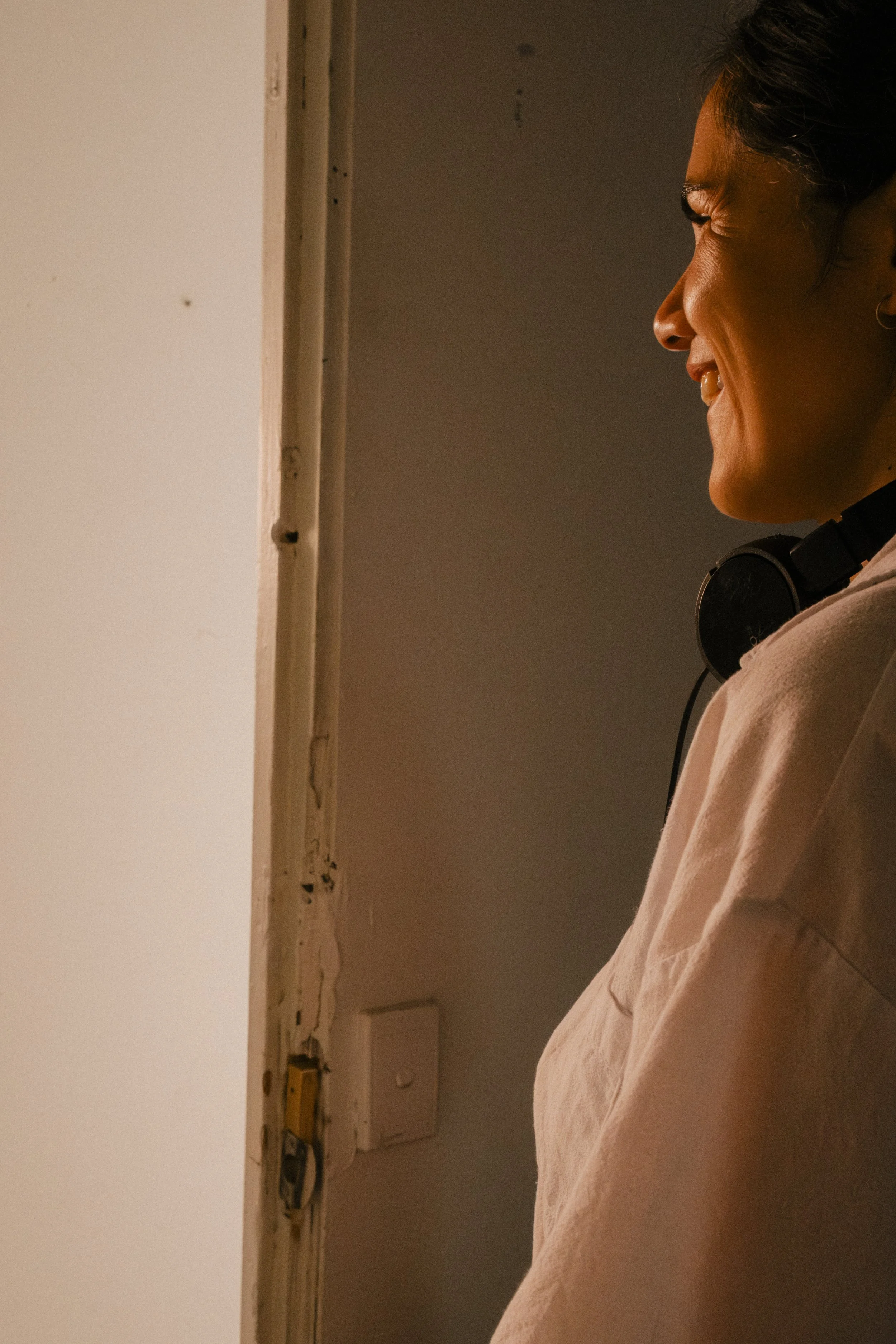 Director watching a scene smiling, wearing headphones around her neck and a light-colored shirt, standing near a doorway with a dark interior behind her, unit still from the short film Stranger Brother.