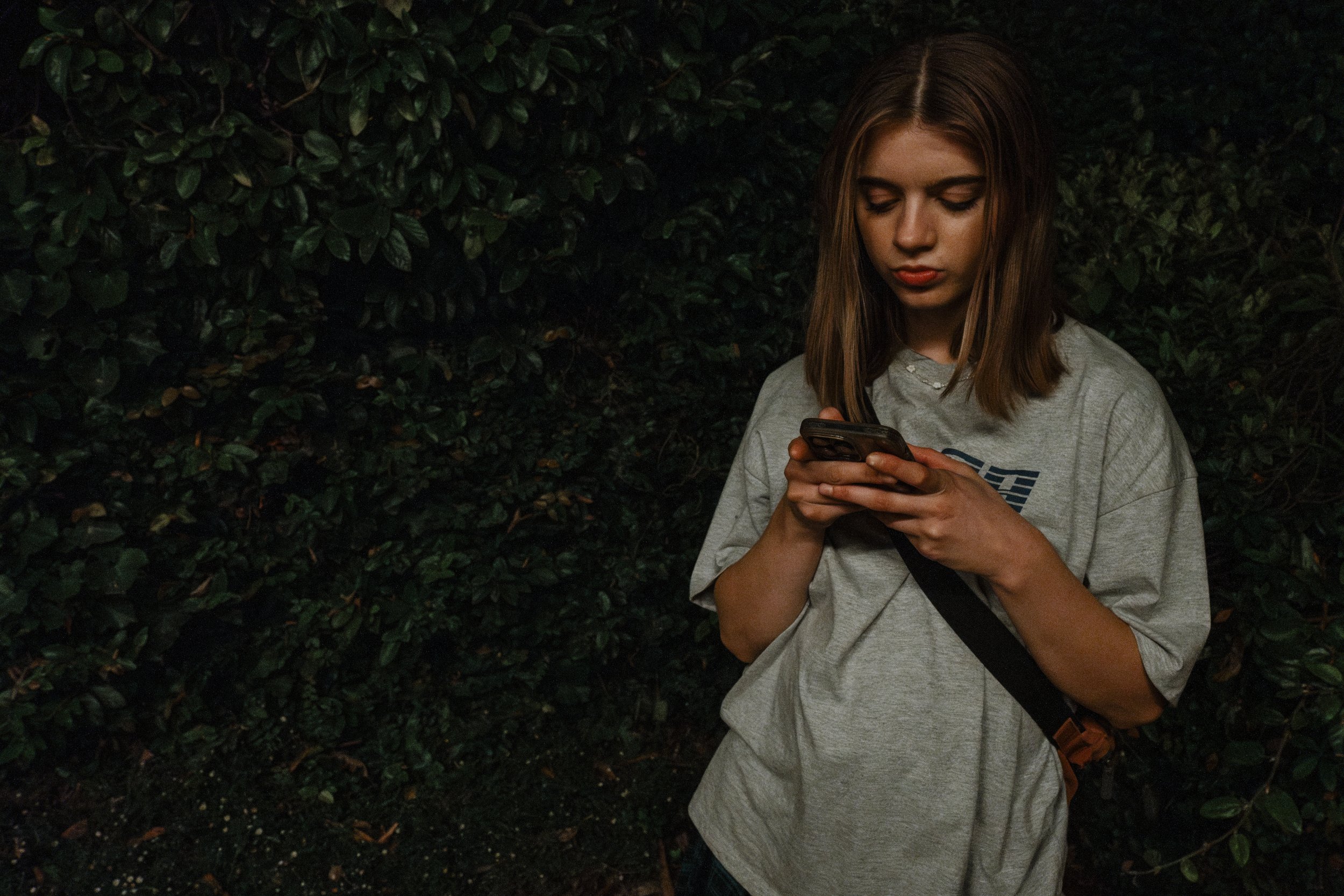 A young woman with shoulder-length brown hair, wearing a gray t-shirt, stands outdoors against a leafy green background, looking at her phone with a neutral expression.