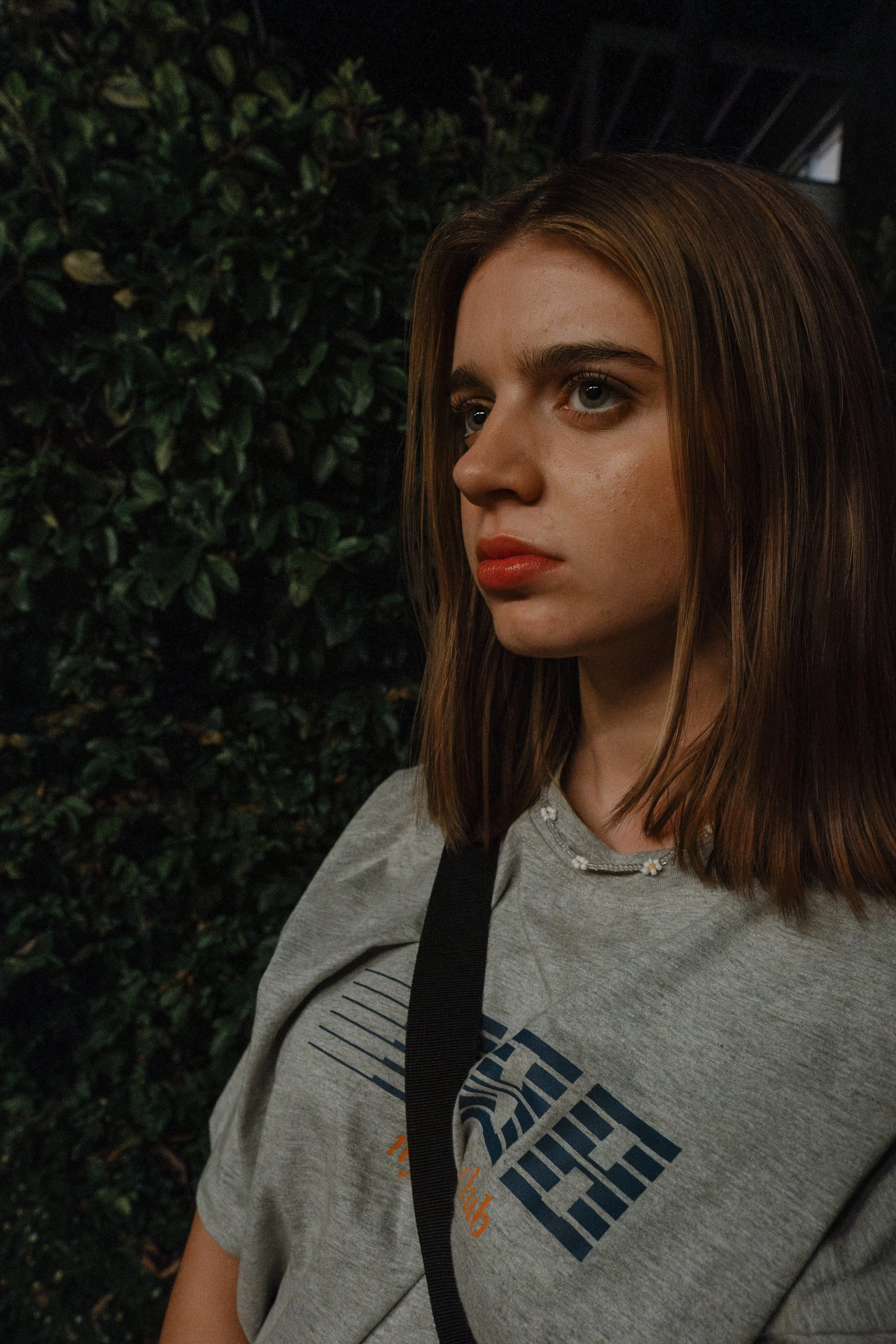 A young woman with medium-length brown hair and blue eyes standing outside near green foliage, wearing a gray t-shirt with a graphic design and a black strap across her shoulder.