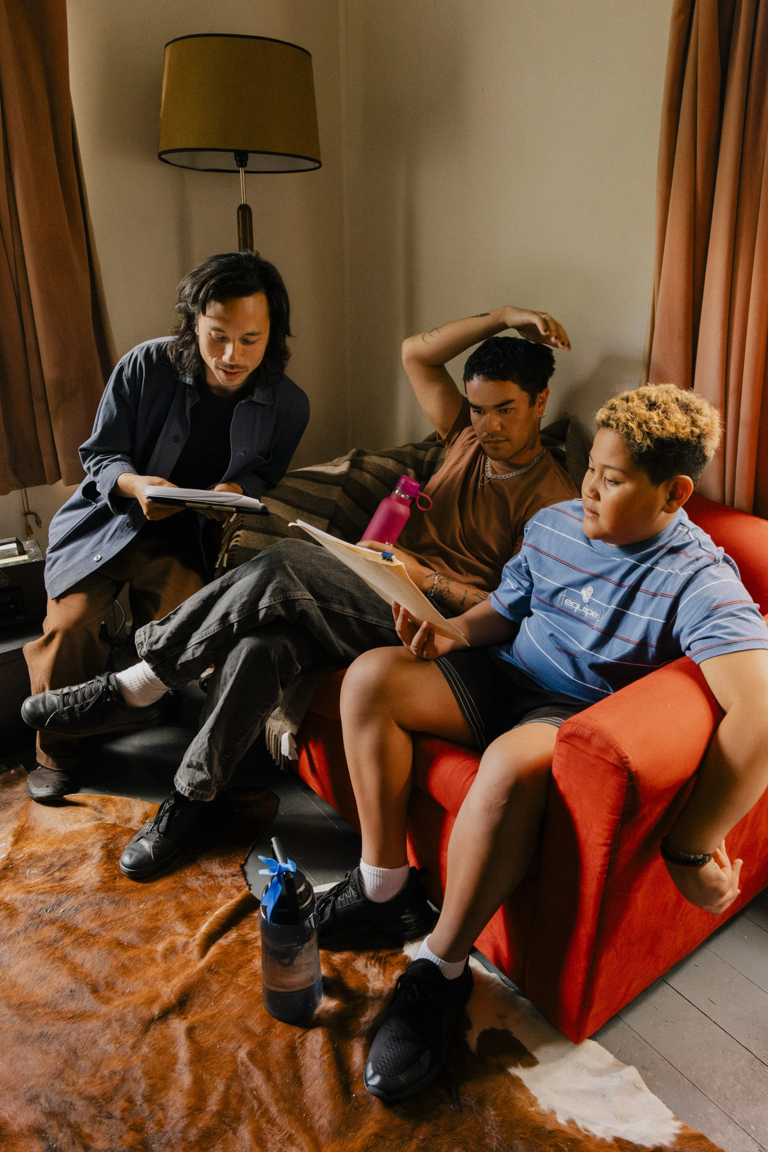 Three young men on a red couch with a brown and black striped blanket, looking at documents, in a room with beige walls, a floor lamp, and brown curtains.