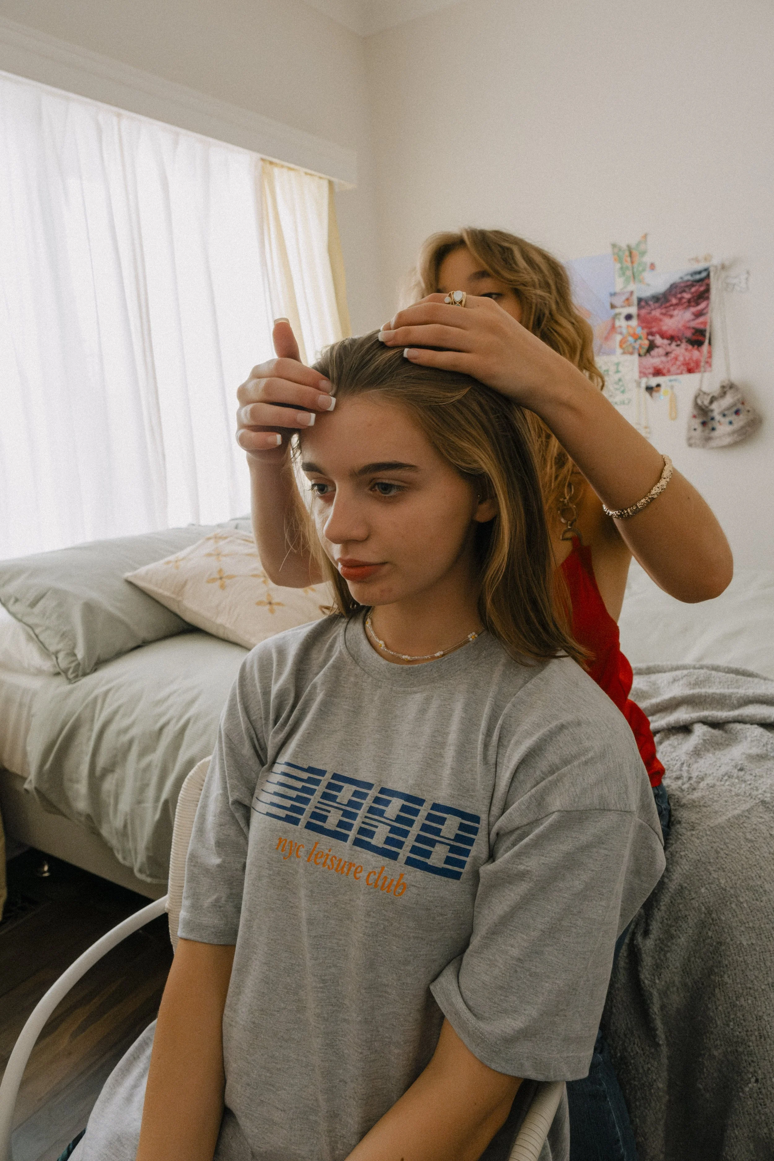 Two women, one standing behind the other, are in a cozy bedroom. The woman in front is seated with her eyes looking down, wearing a gray T-shirt that reads "NYC leisure club." The woman behind is touching the seated woman's hair and face, appearing to style or examine her hair.