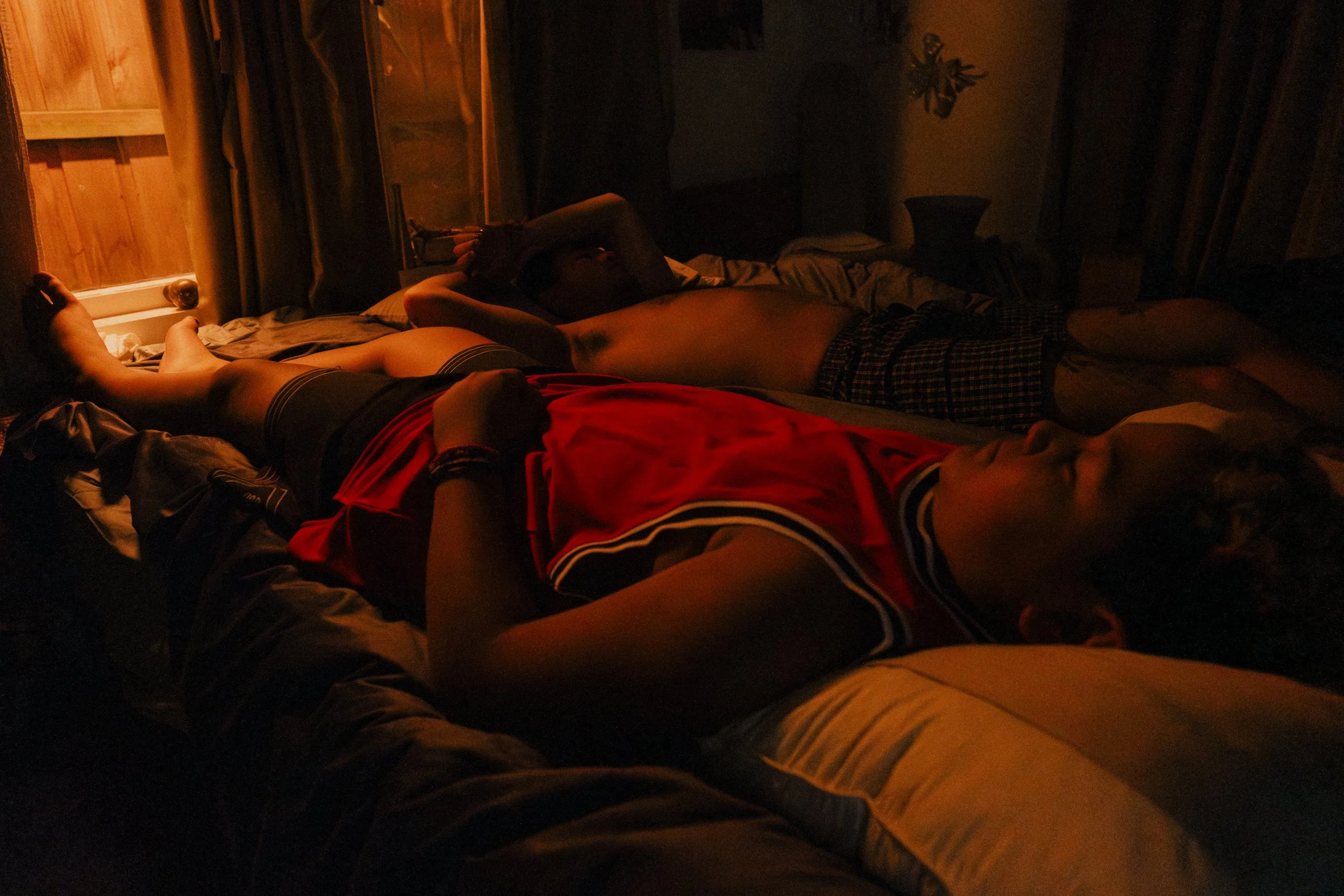 Two young men lying on beds in a dimly lit room, one wearing a red sports jersey and the other shirtless, with beds and curtains visible in the background, unit still from the short film Stranger Brother.