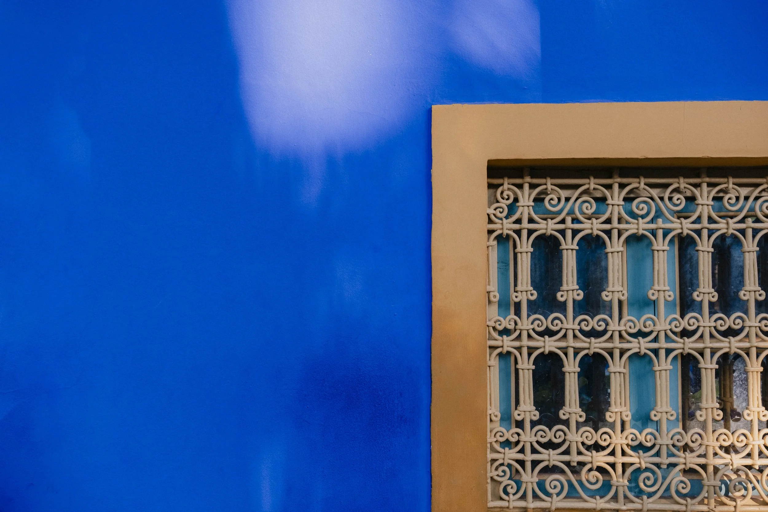 Close-up of a beige decorative iron window grille on a vibrant blue wall with a beige window frame.