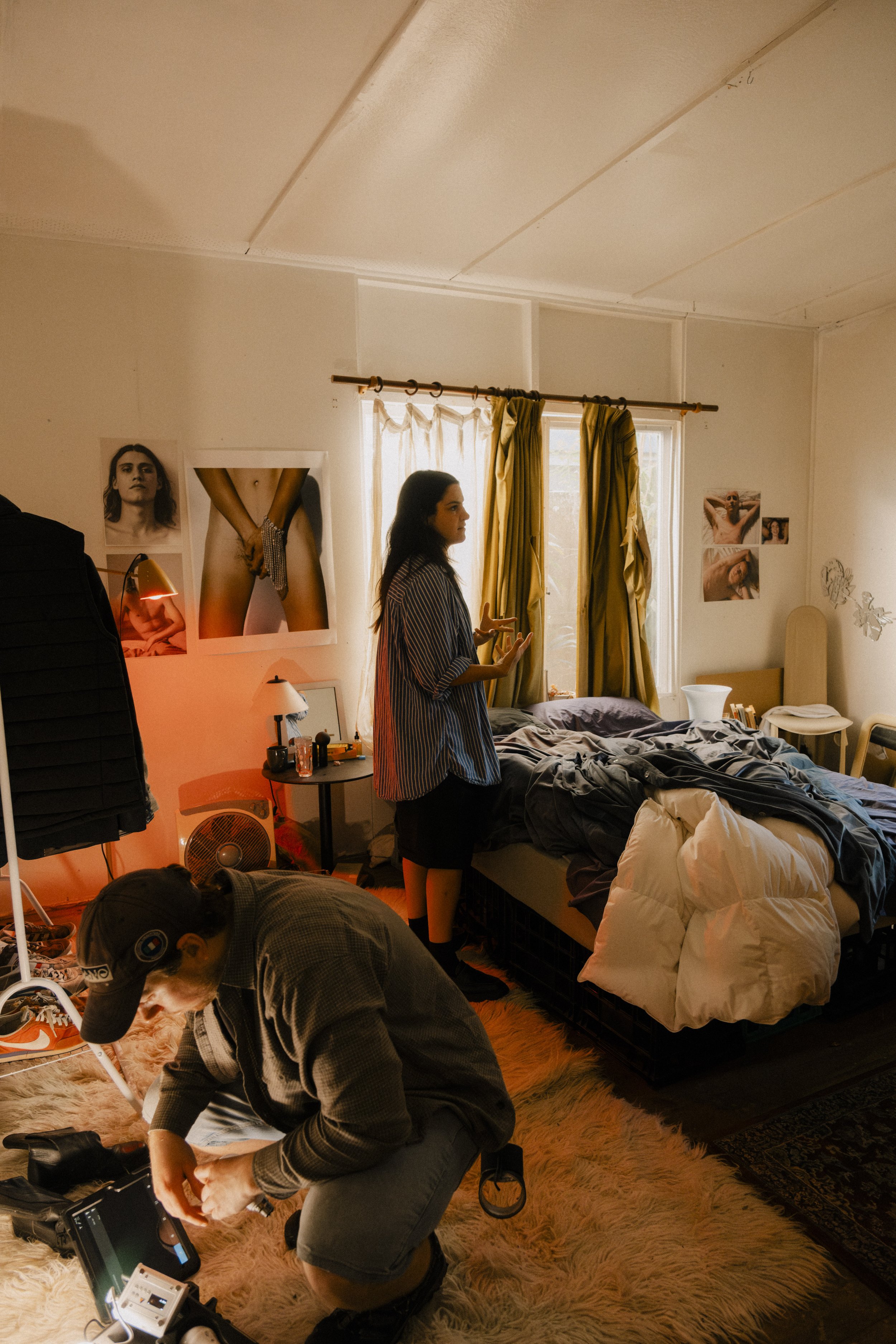 A woman standing next to a bed covered with clothes and blankets, talking, with sunlight coming through a window, while a man kneels on the floor working with camera equipment in a cluttered bedroom, unit still from the short film Stranger Brother.