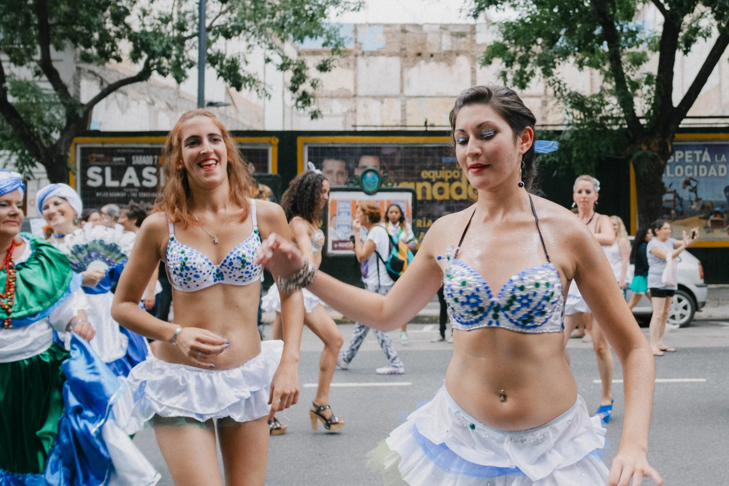 Women dancing in colorful costumes on a street during a parade, with onlookers and billboards in the background.