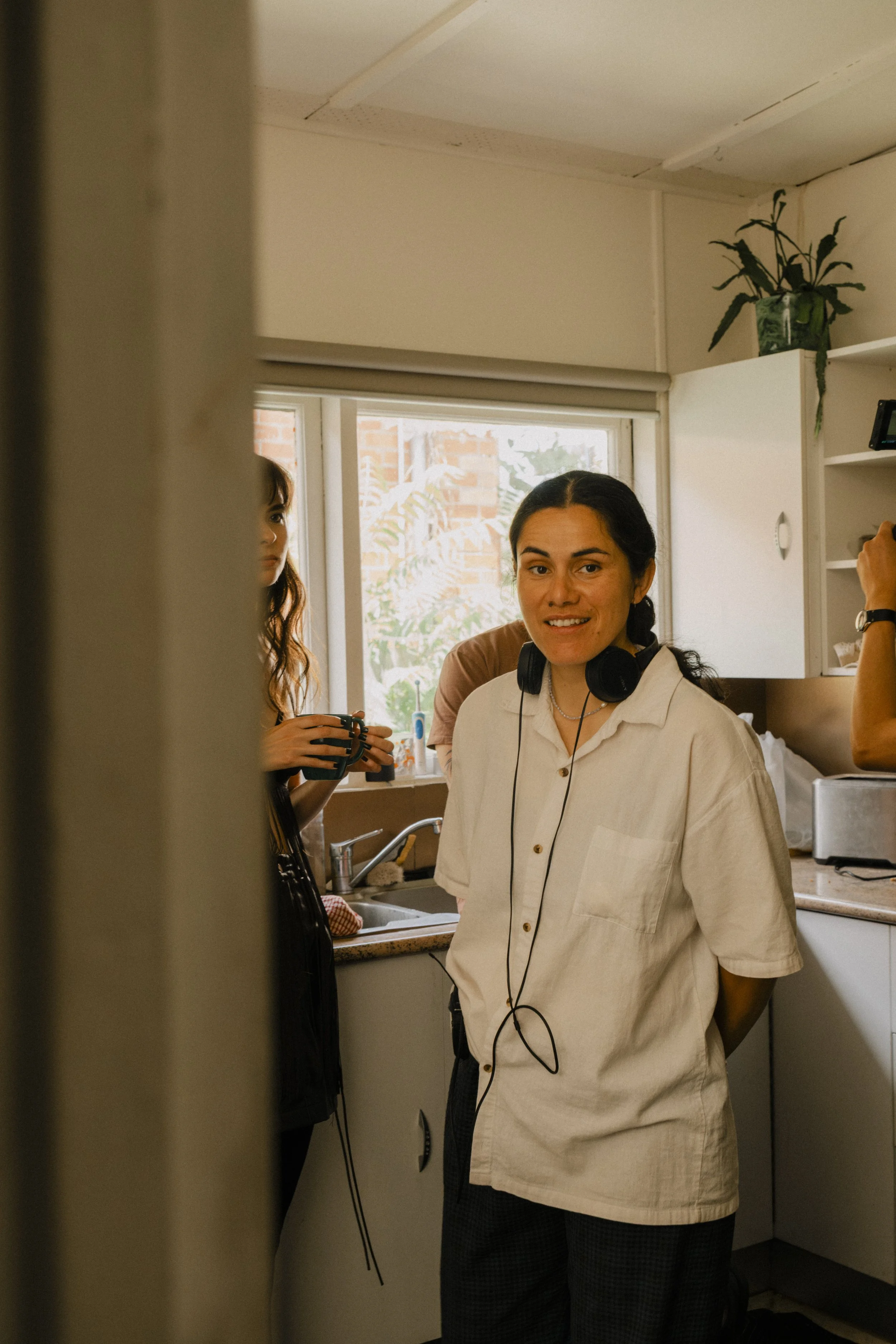 A woman with dark hair in a braid, wearing a white shirt with headphones around her neck, smiling at the camera in a kitchen with two other people in the background.