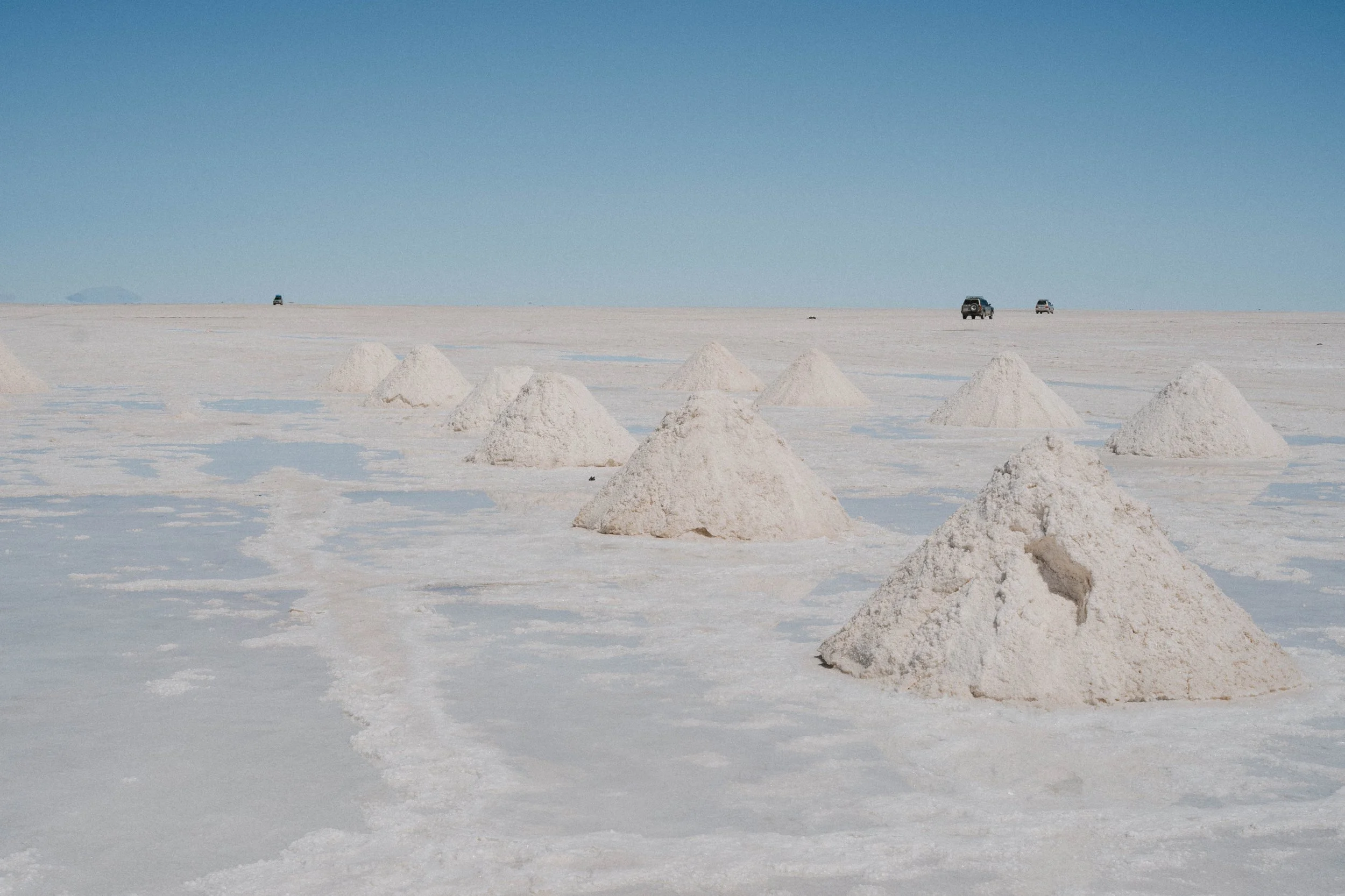 Multiple conical salt piles in the salt flats with a clear blue sky and a few vehicles in the distance.