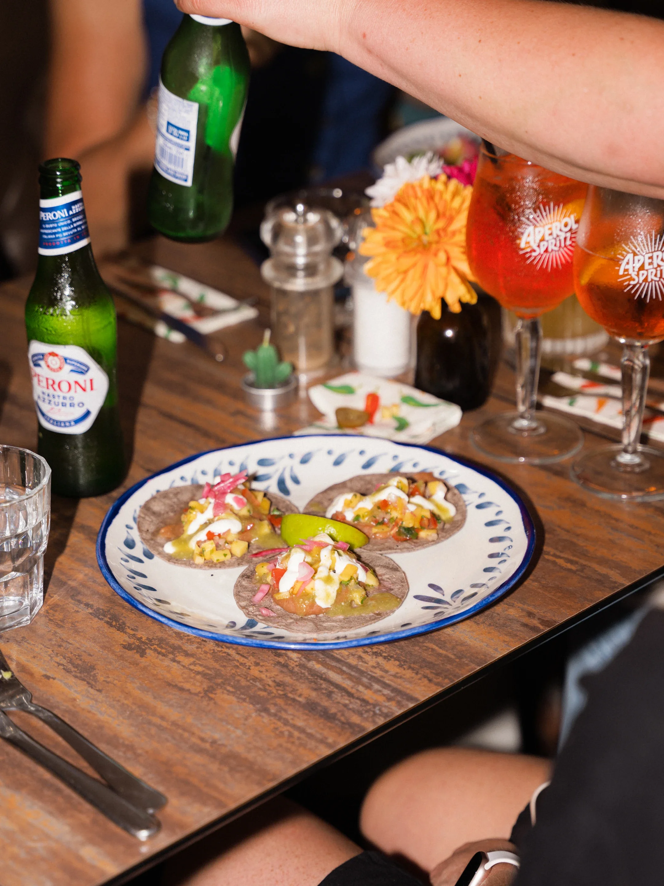 Plate of Mexican-style tacos with toppings beside Peroni beer bottles and Aperol Spritz cocktails on a restaurant table
