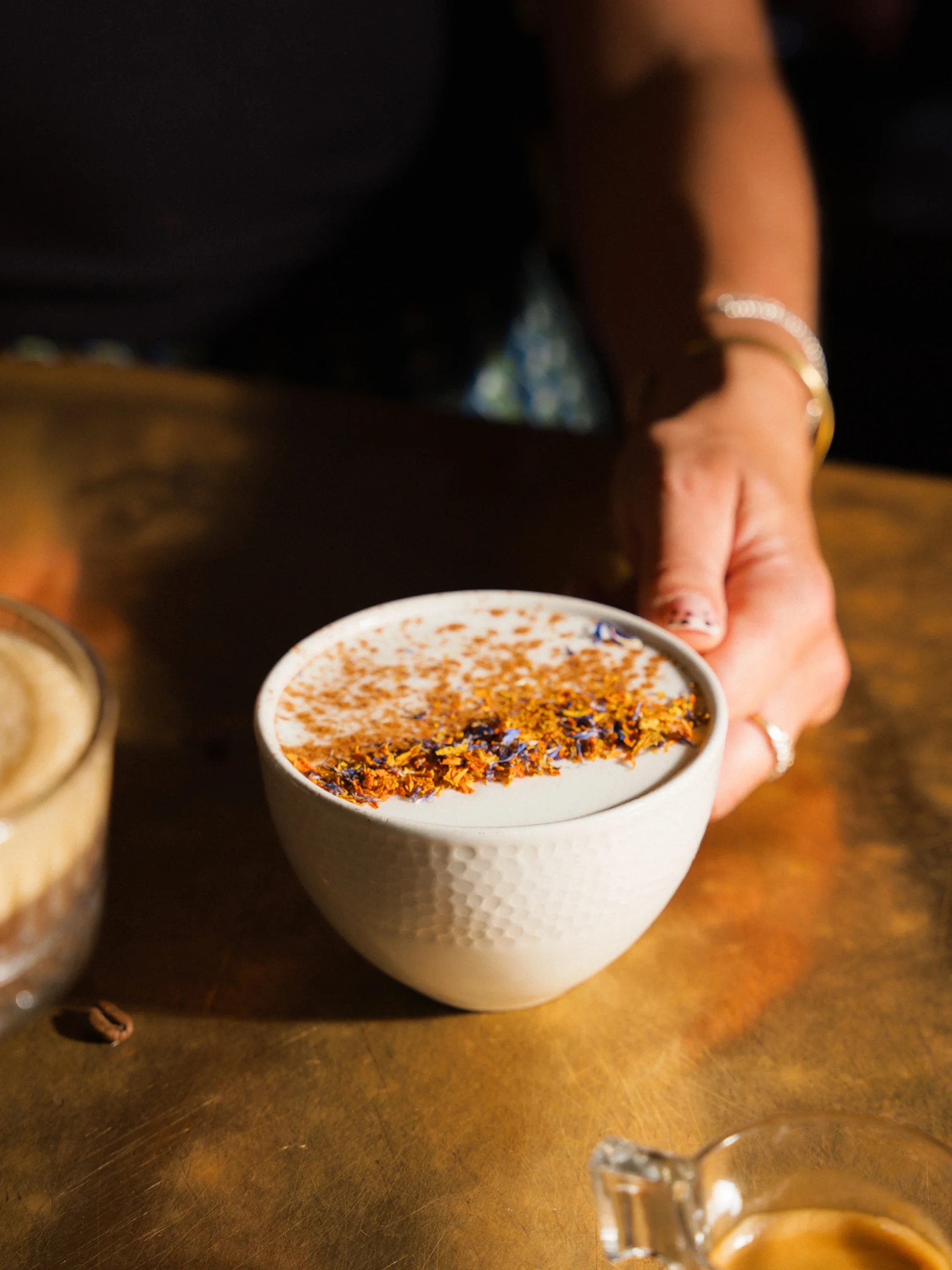A hand holds a white bowl of latte art coffee topped with dried orange and purple flower petals on a wooden table.