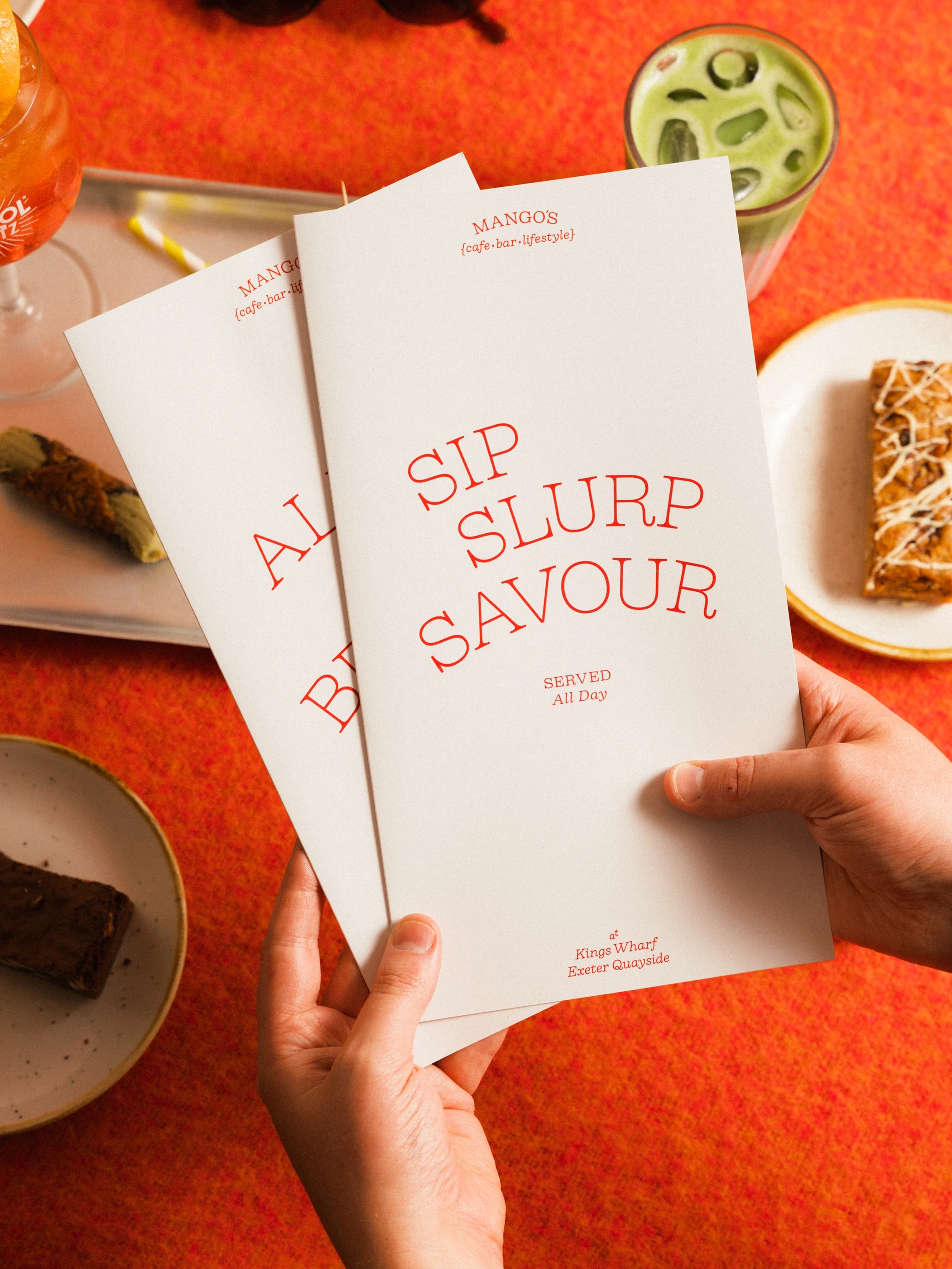 Hands holding a Mango's cafe menu reading “Sip, Slurp, Savor” beside drinks and plated food.