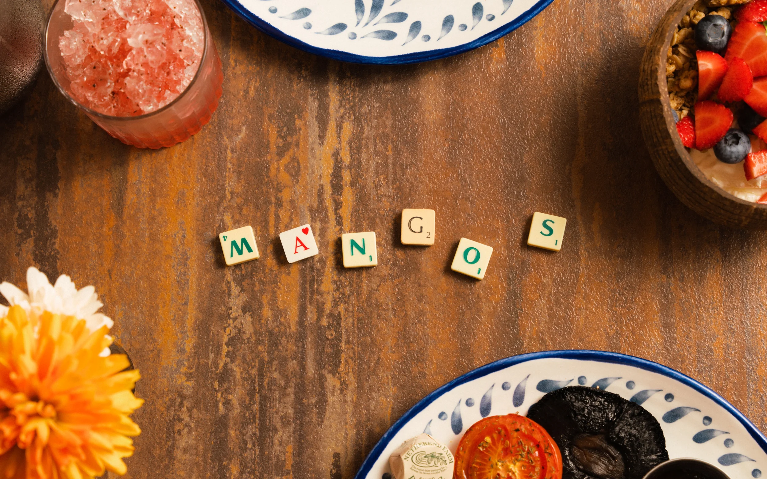 Overhead view of a rustic table with Scrabble tiles spelling “WANDS,” a pink drink, berry granola bowl, plates of food, and an orange flower.