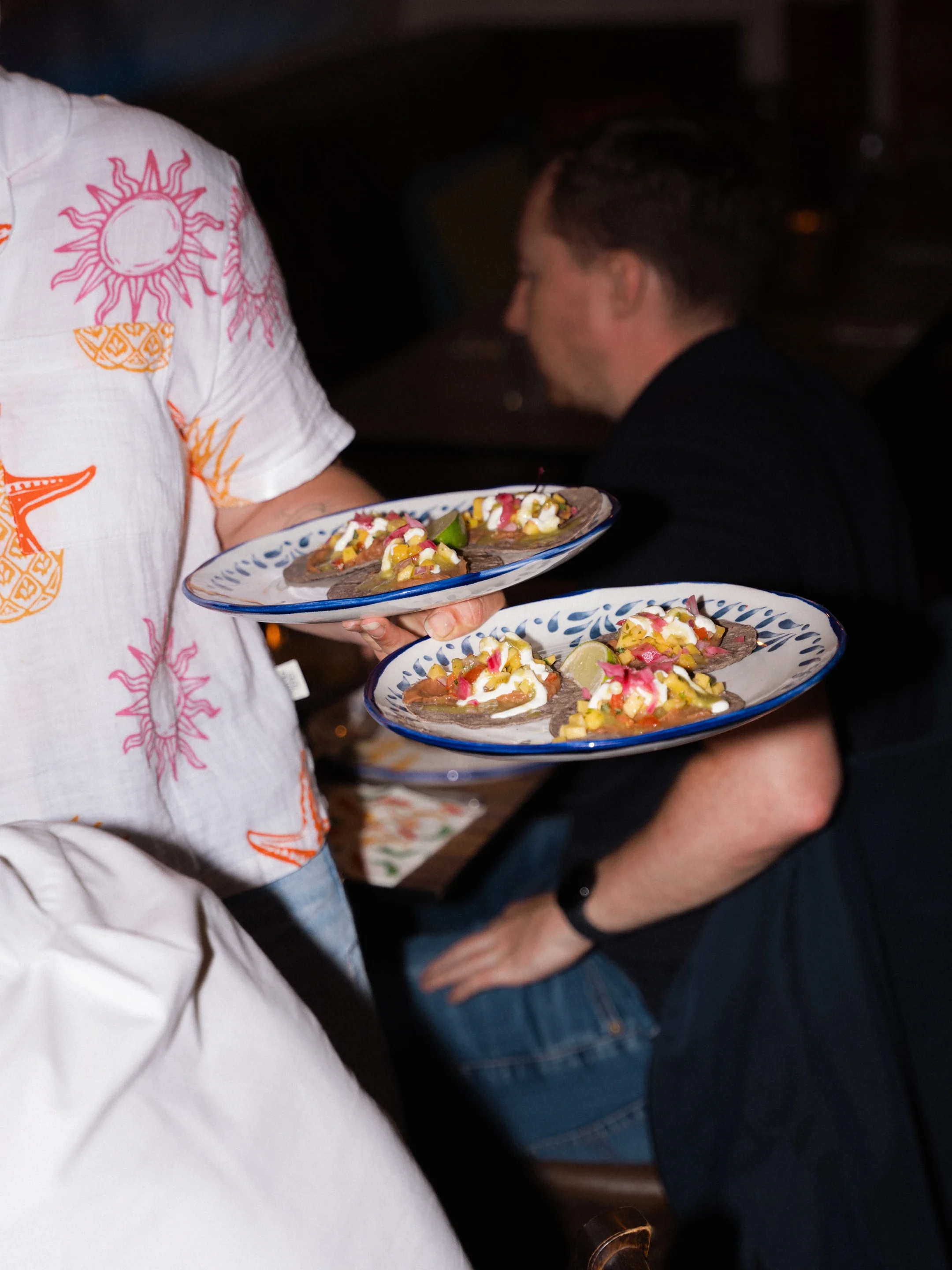 Two people carry plates of colorful tostadas topped with salsa, crema, vegetables, and lime at a dimly lit restaurant.